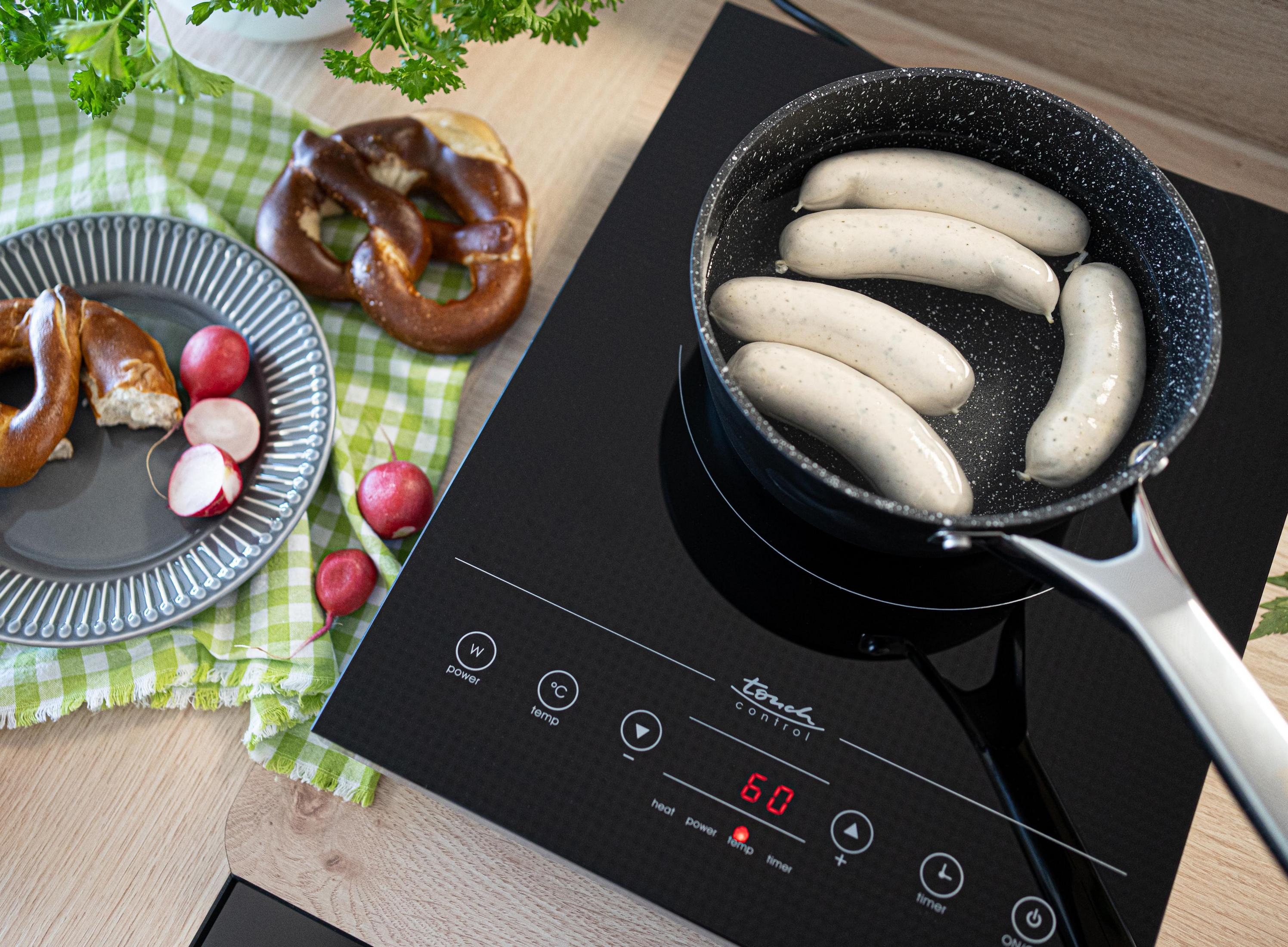 Płyta indukcyjna z kiełbasą w patelni, preclami i rzodkiewkami na kraciastej ściereczce. Przygotowywanie jedzenia.