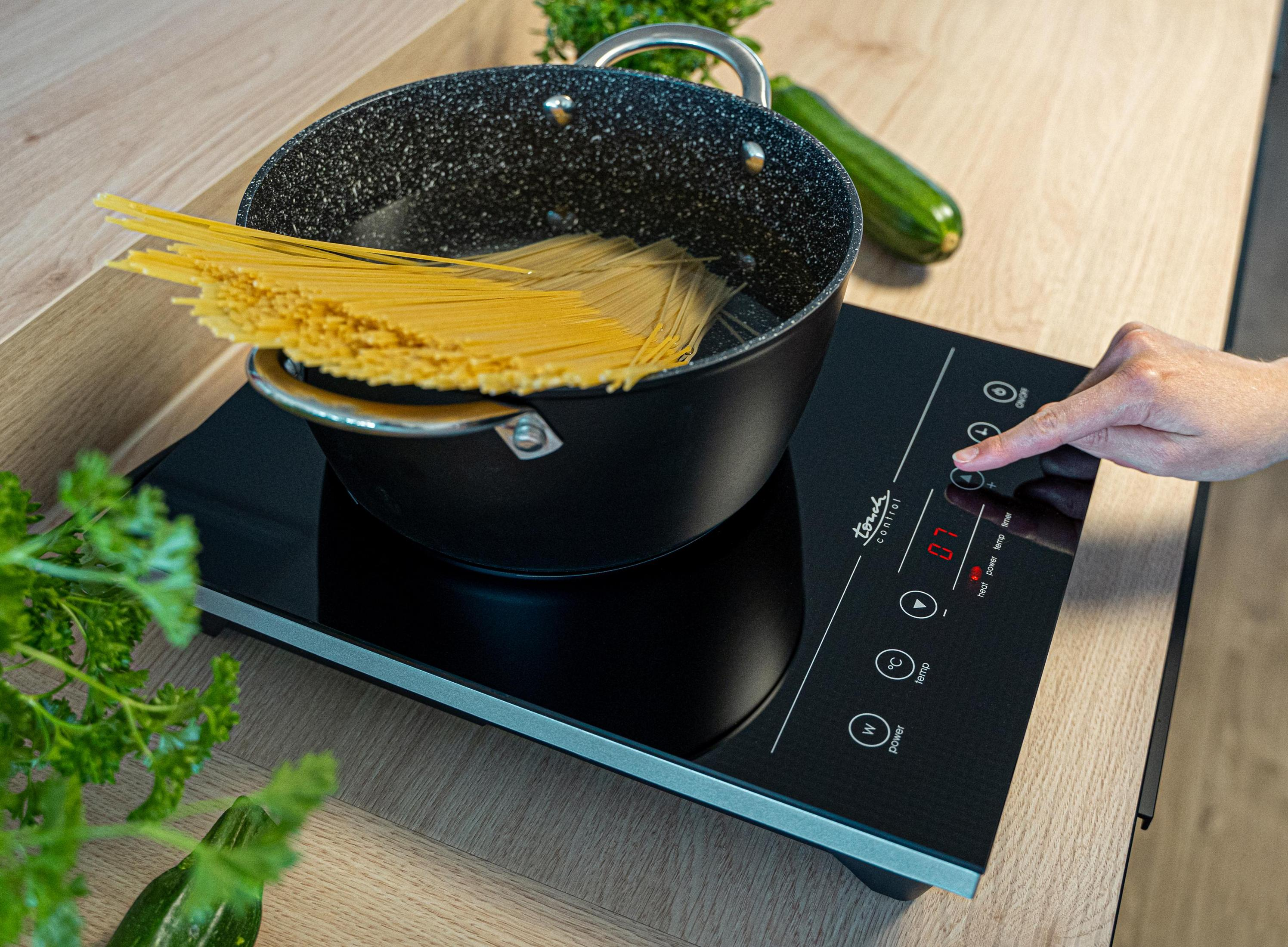 Scena gotowania: Spaghetti w garnku na płycie indukcyjnej, dłoń dotyka panelu sterowania.