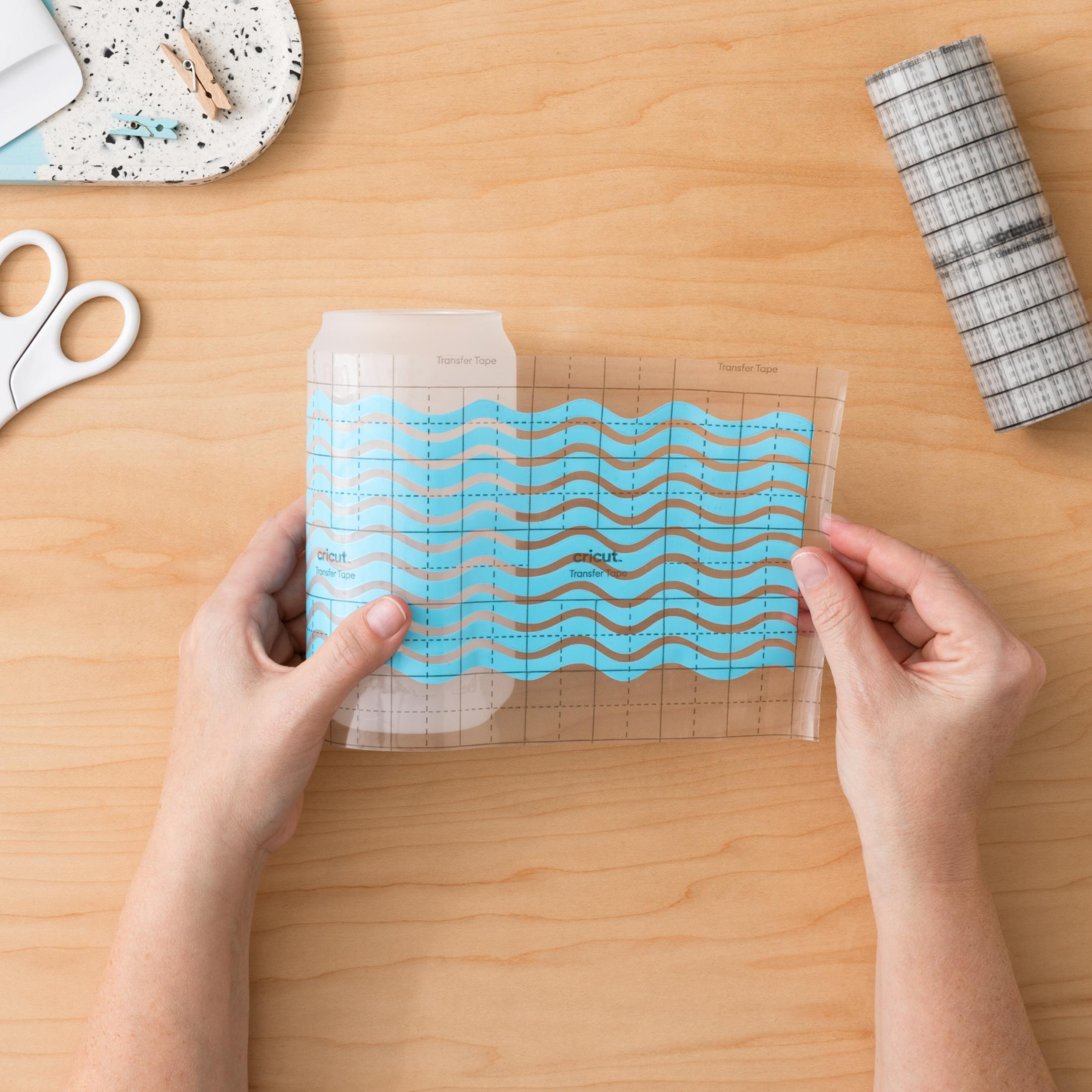 Hände, die Cricut-Transferband über einem blauen Wellendesign auf einem hellen Holztisch halten.