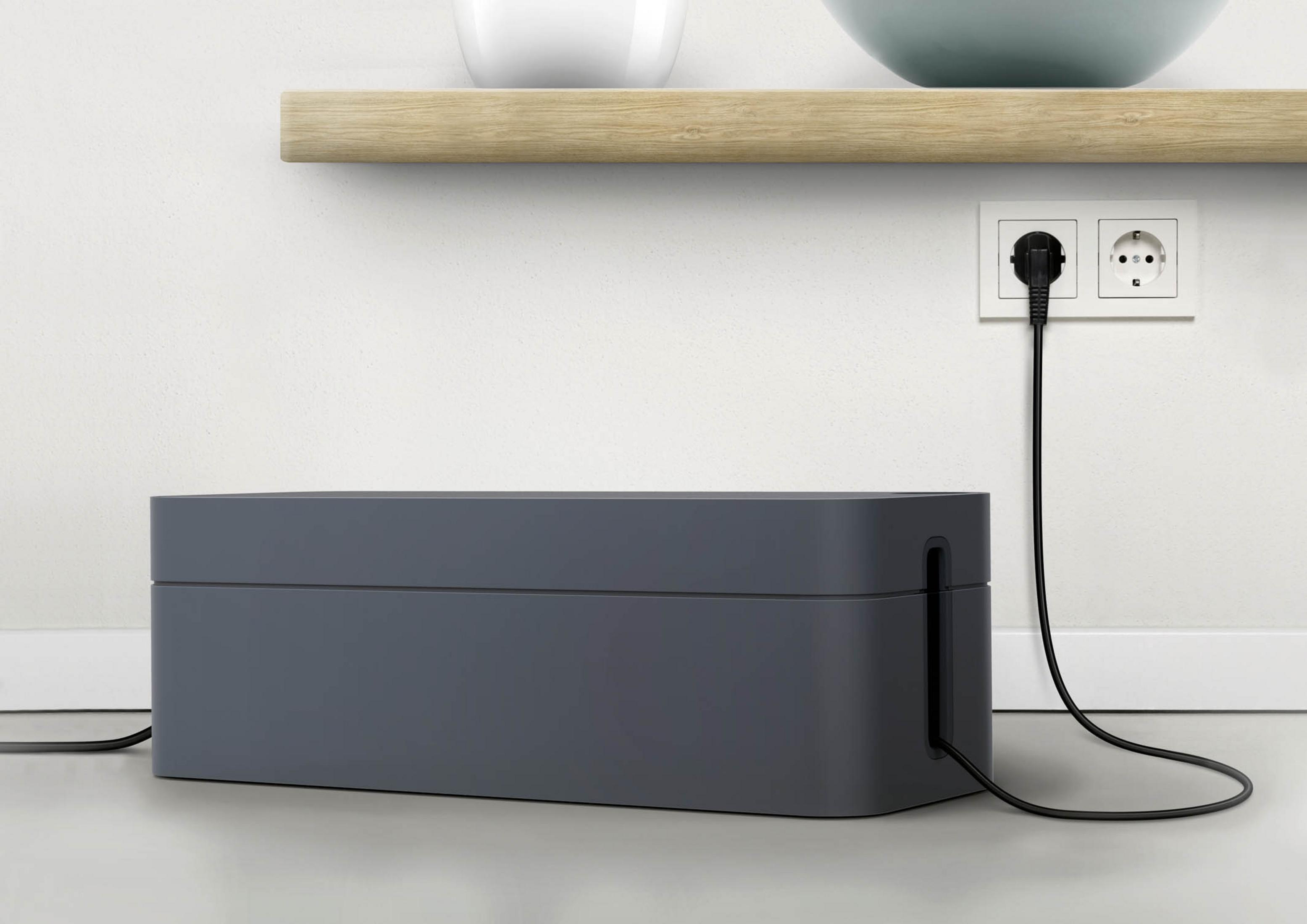 Dunkelgraue Kabelbox. Kabel kommen heraus. Weiße Wand mit Steckdose. Holzregal darüber. Weißer Boden.