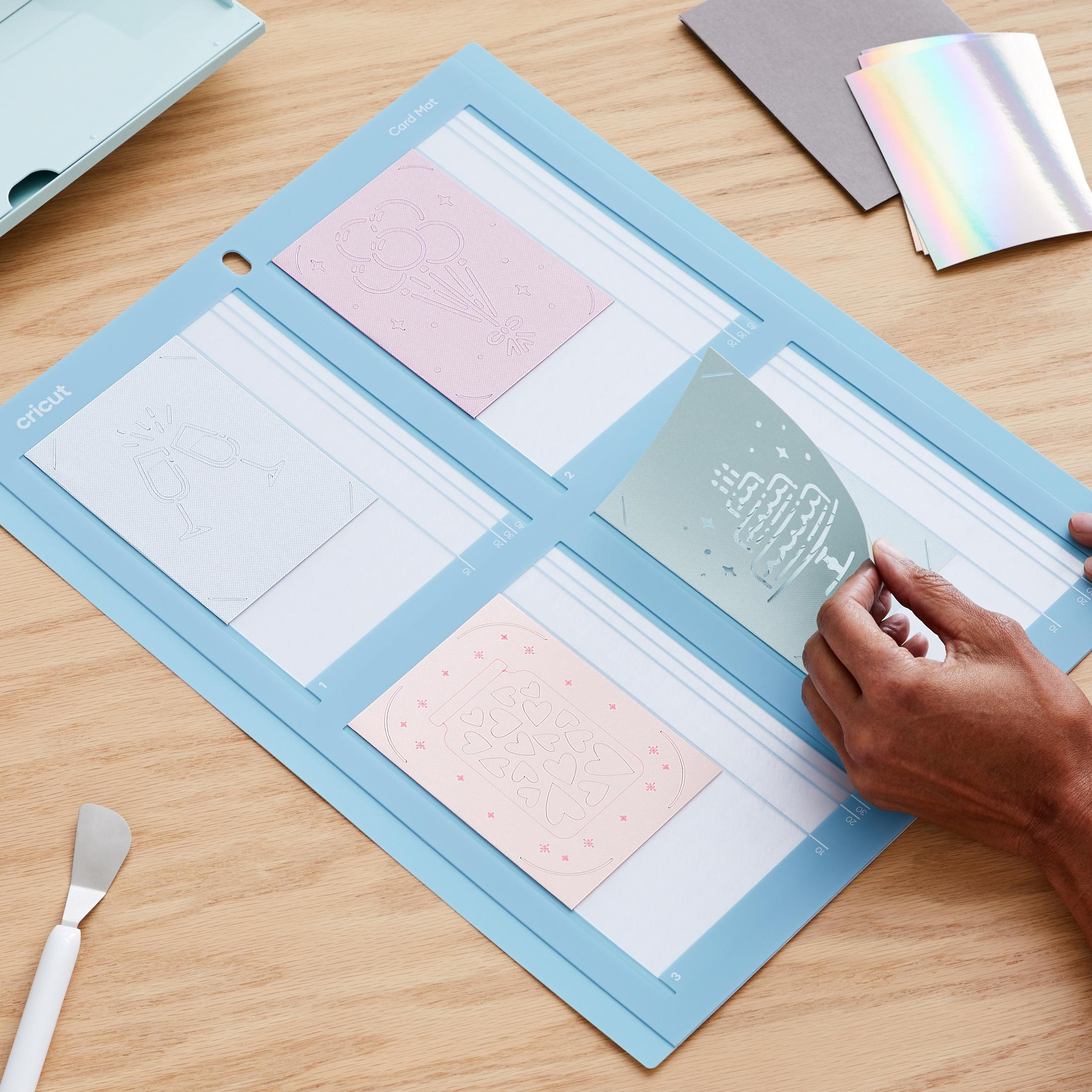 Person verwendet Cricut-Maschine zum Schneiden von Karten. Blaue Schneidematte, mit geschnittenen Karten. Auf einem Holztisch.