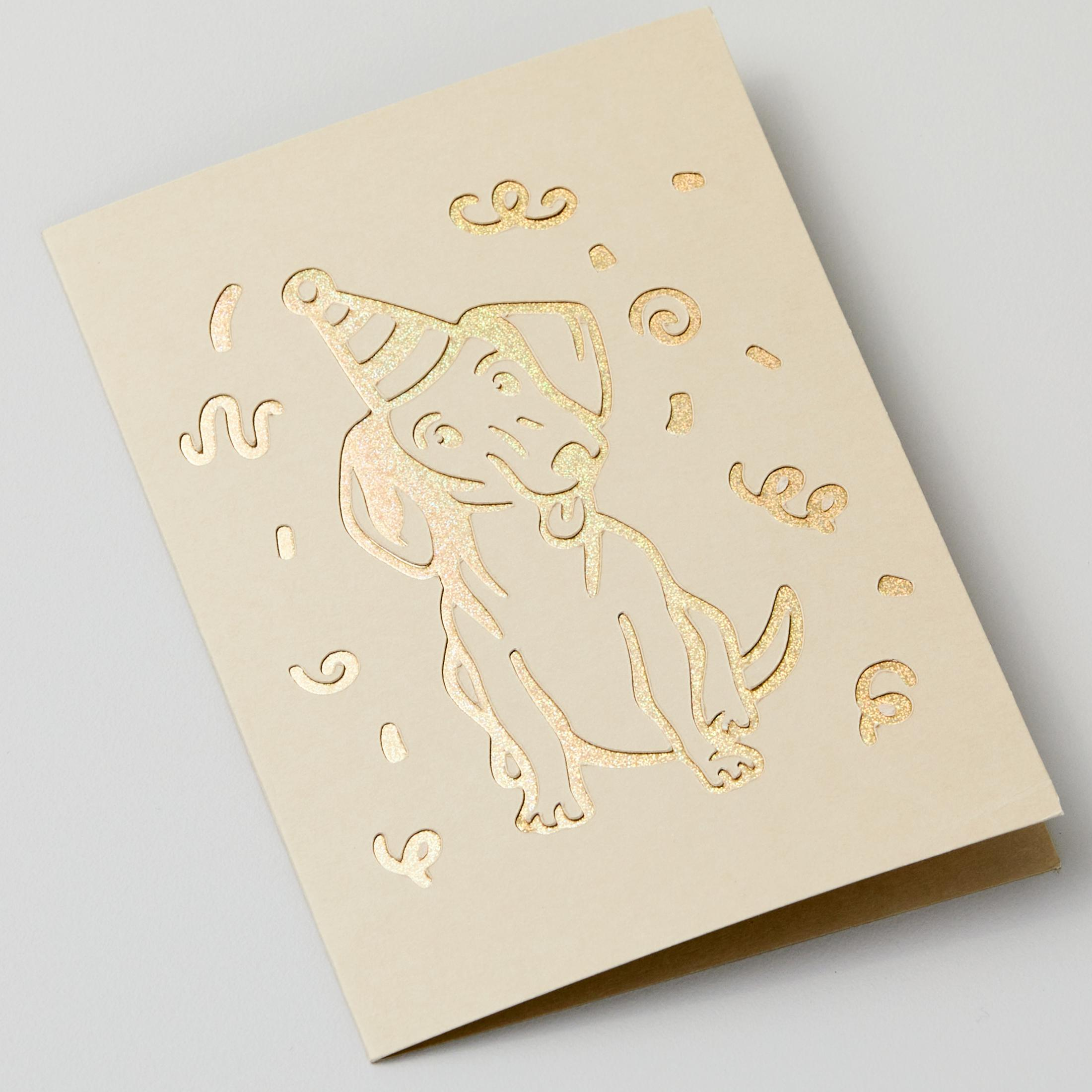 Cremefarbene Grußkarte mit Hundeillustration. Sie hat eine Partyhut- und Konfetti-Ausschnitte.