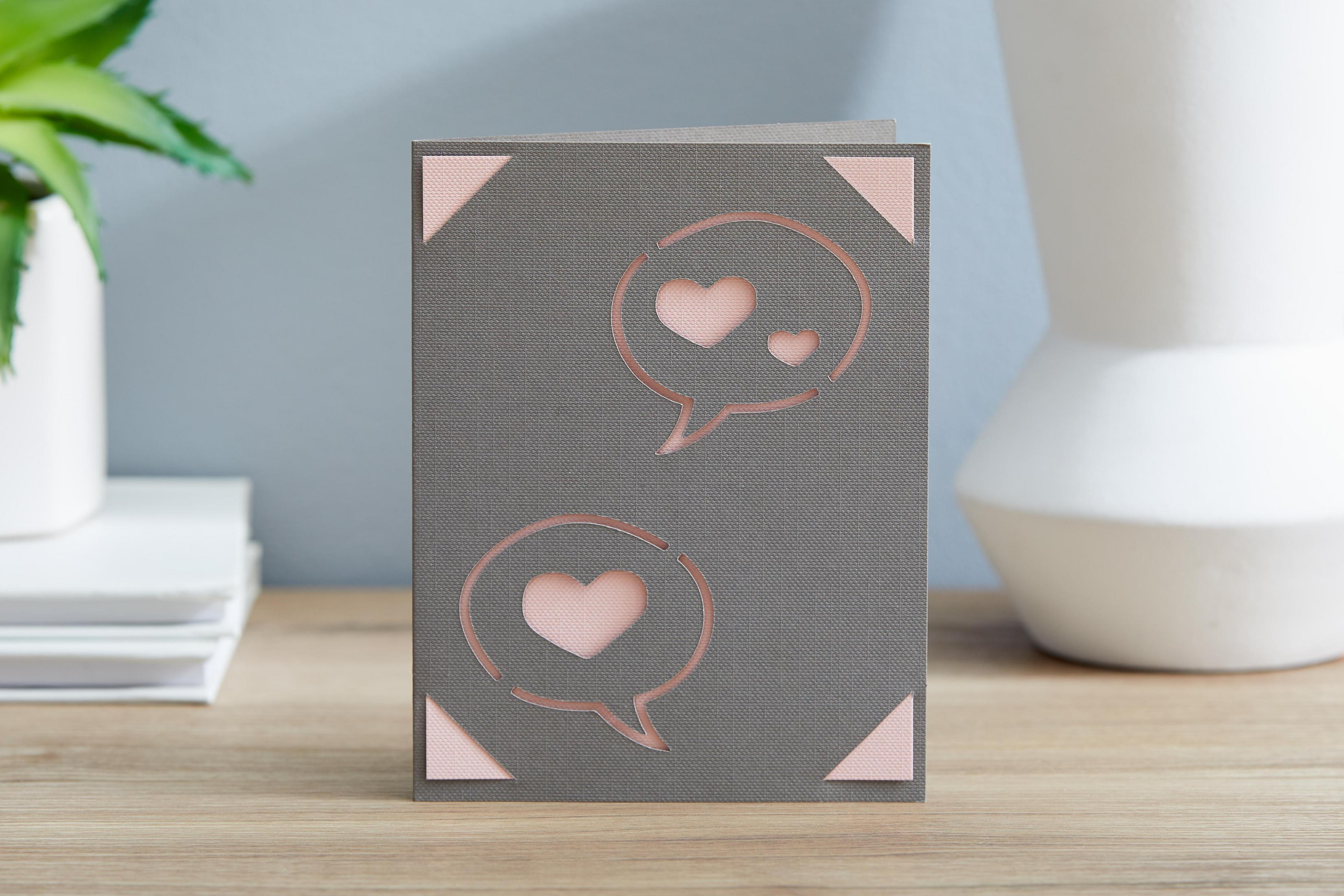 Eine Karte mit ausgeschnittenen Sprechblasen mit Liebesherzen. Die Karte ist grau mit rosa Akzenten.