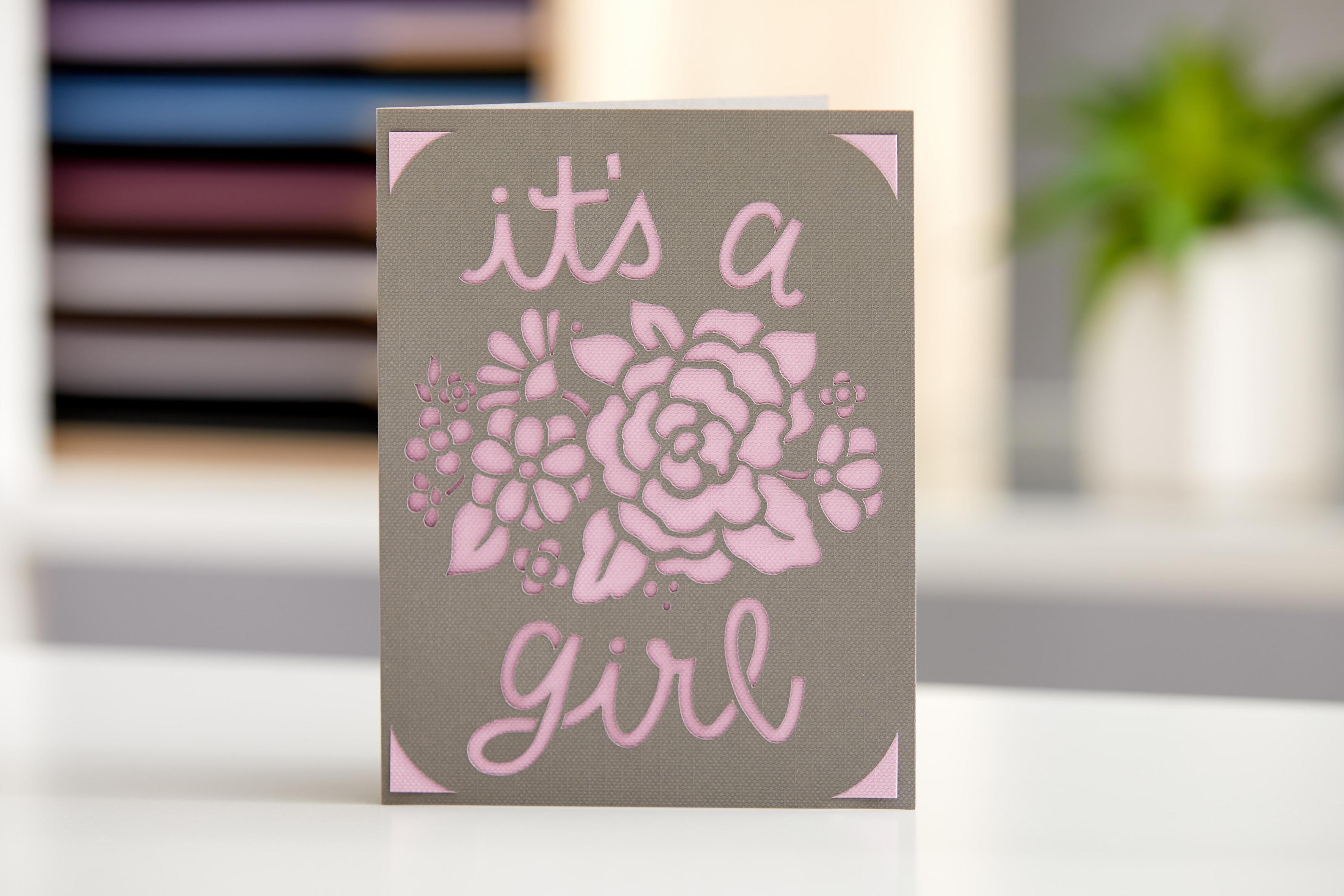 Eine graue Karte mit einem rosa Blumendesign und den Worten 'it's a girl' in rosa Buchstaben.