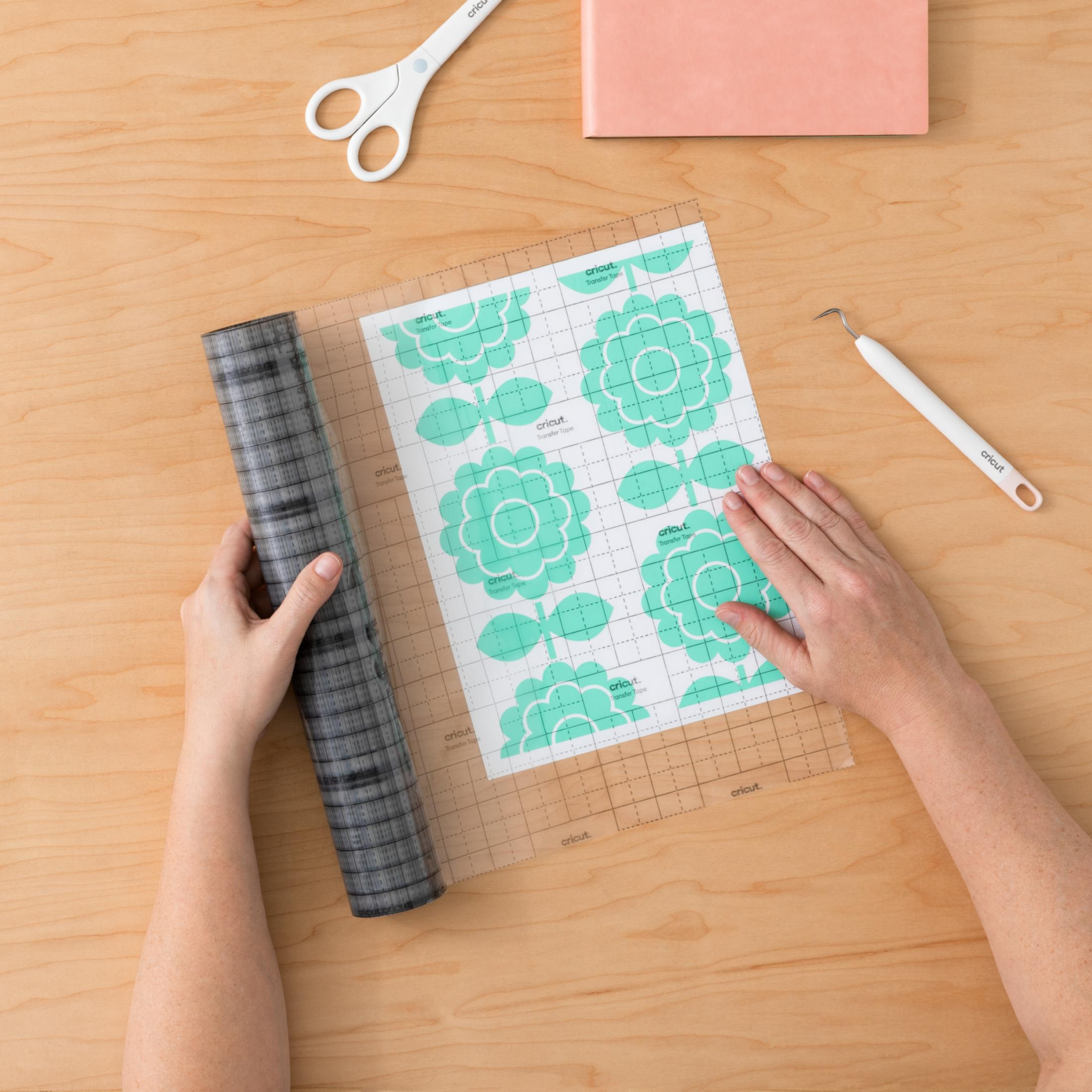 Hände, die mit Cricut-Materialien arbeiten. Es gibt eine Rolle, eine Matte mit Blumen, eine Schere und einen Haken.