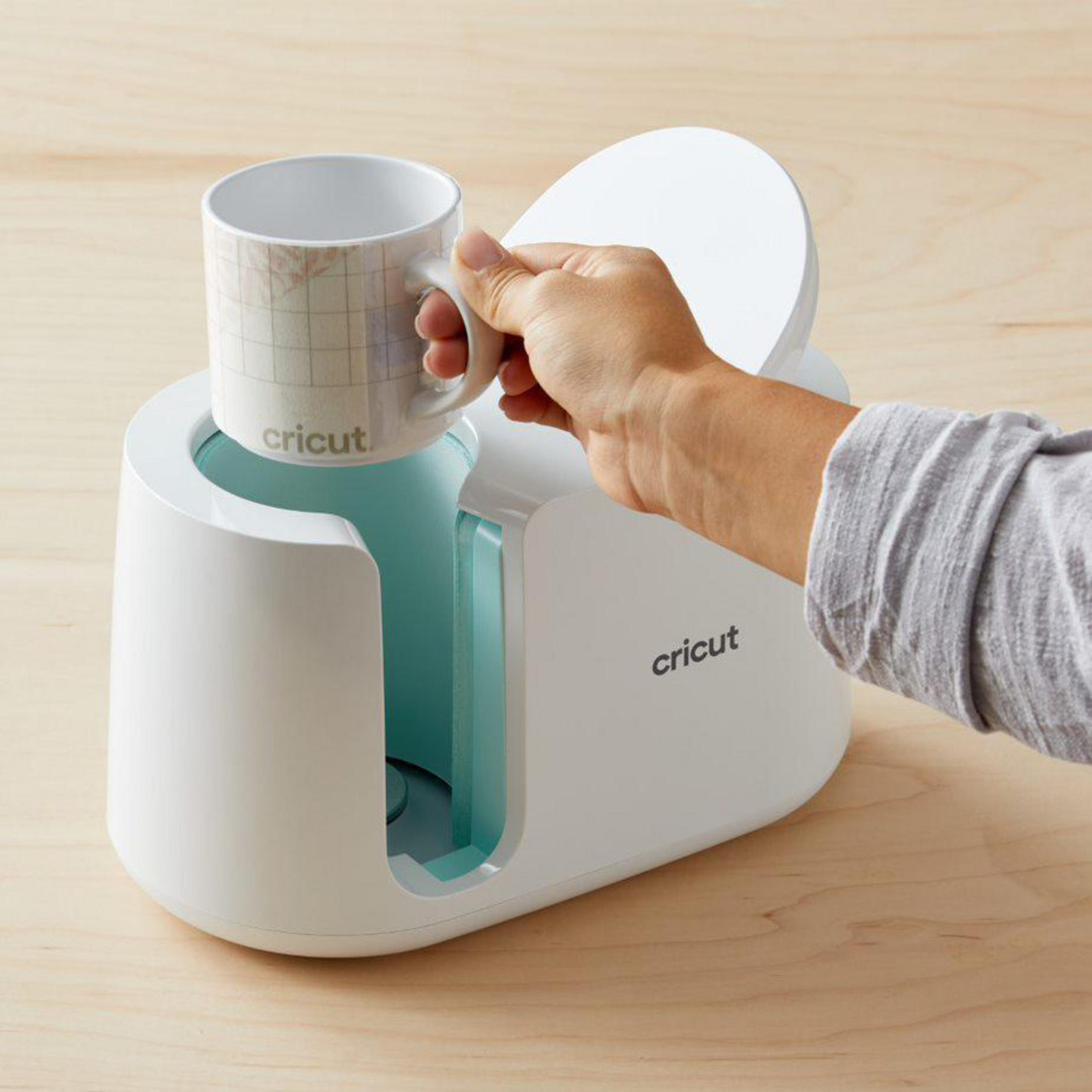 Person hält einen Becher in einer weißen Cricut-Becherpresse auf einer hellen Holzoberfläche.
