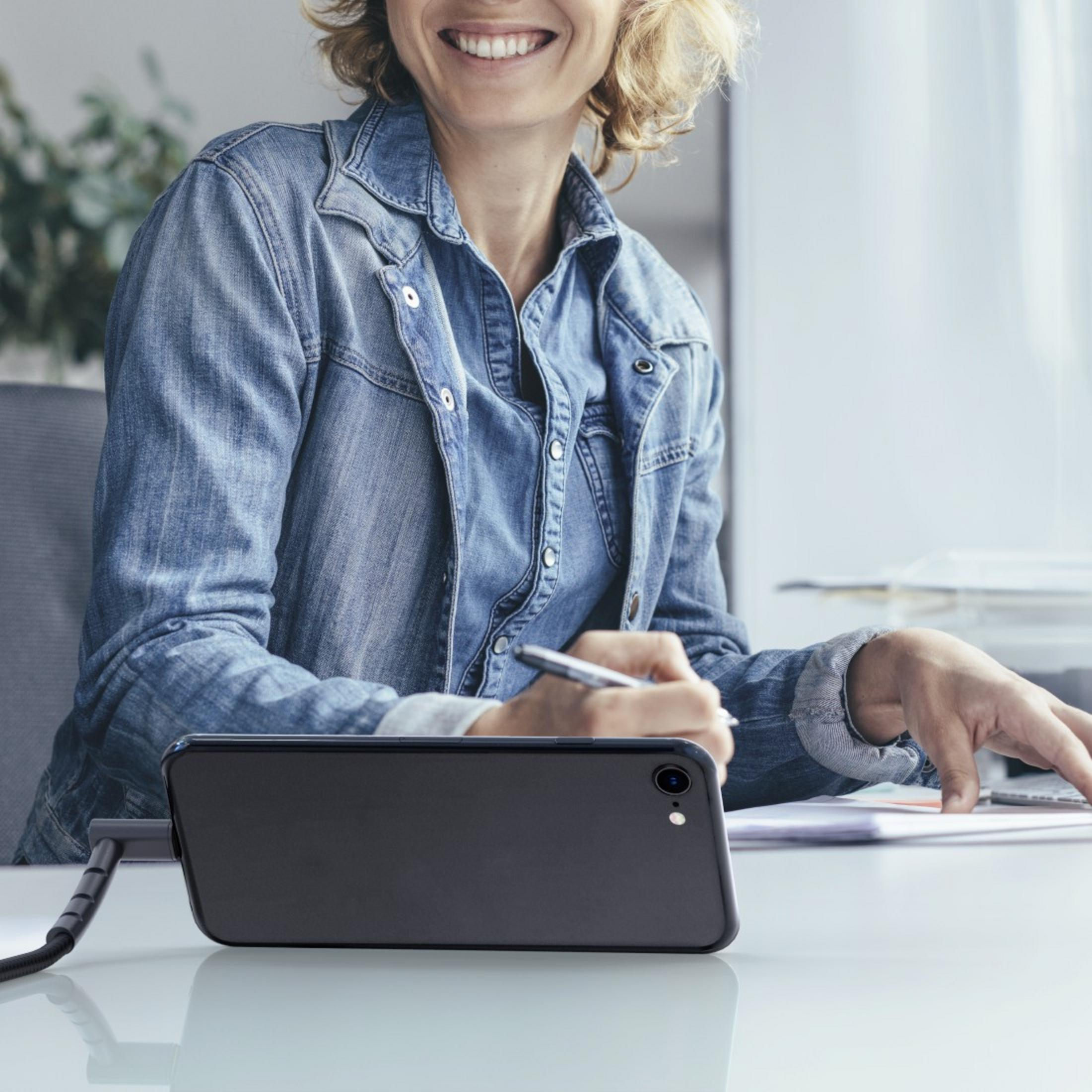 Frau in Jeansjacke arbeitet an einem Schreibtisch mit einem an ein Kabel angeschlossenen Telefon.