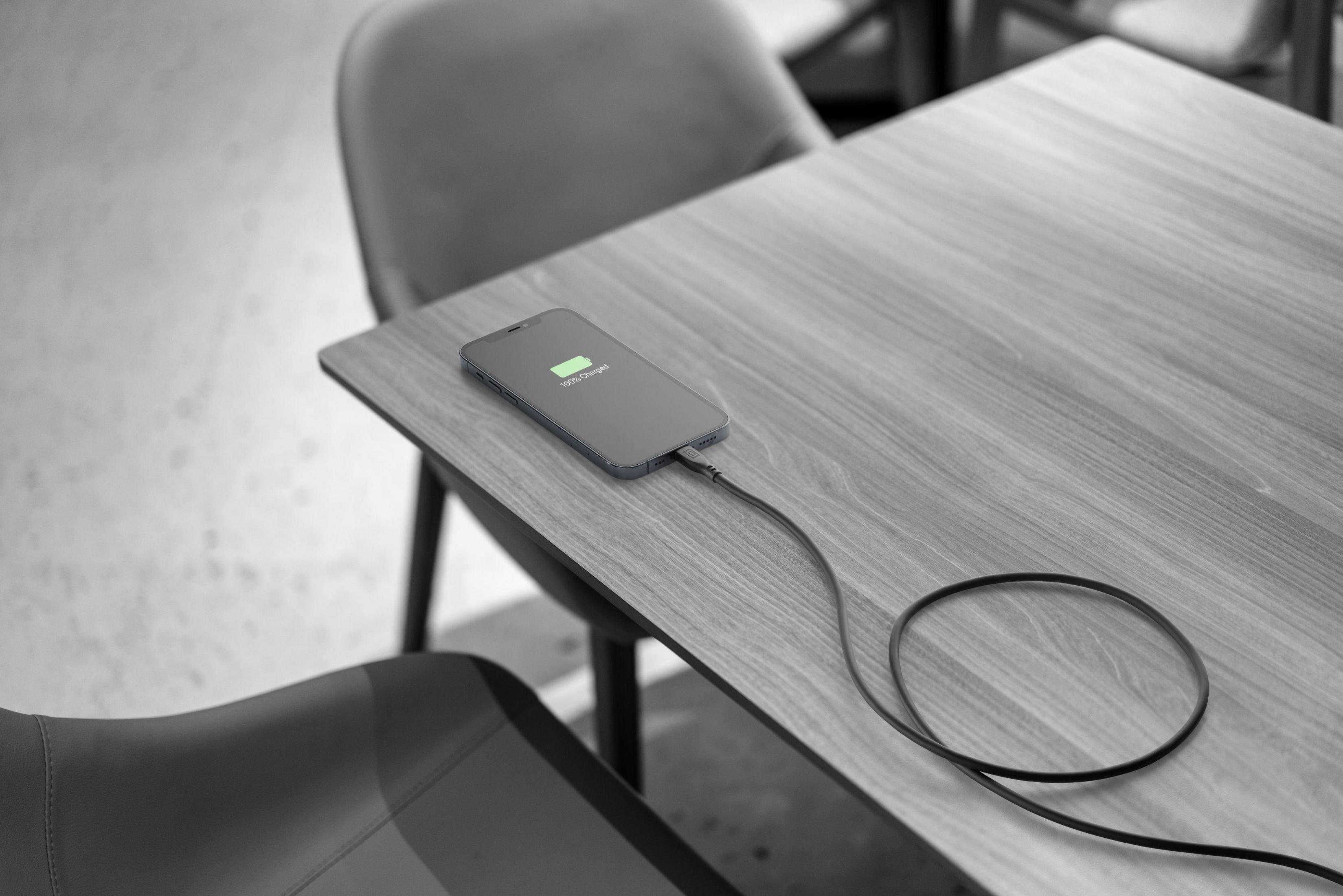 Image en niveaux de gris d'un téléphone en charge sur une table en bois, avec câble noir et chaises.