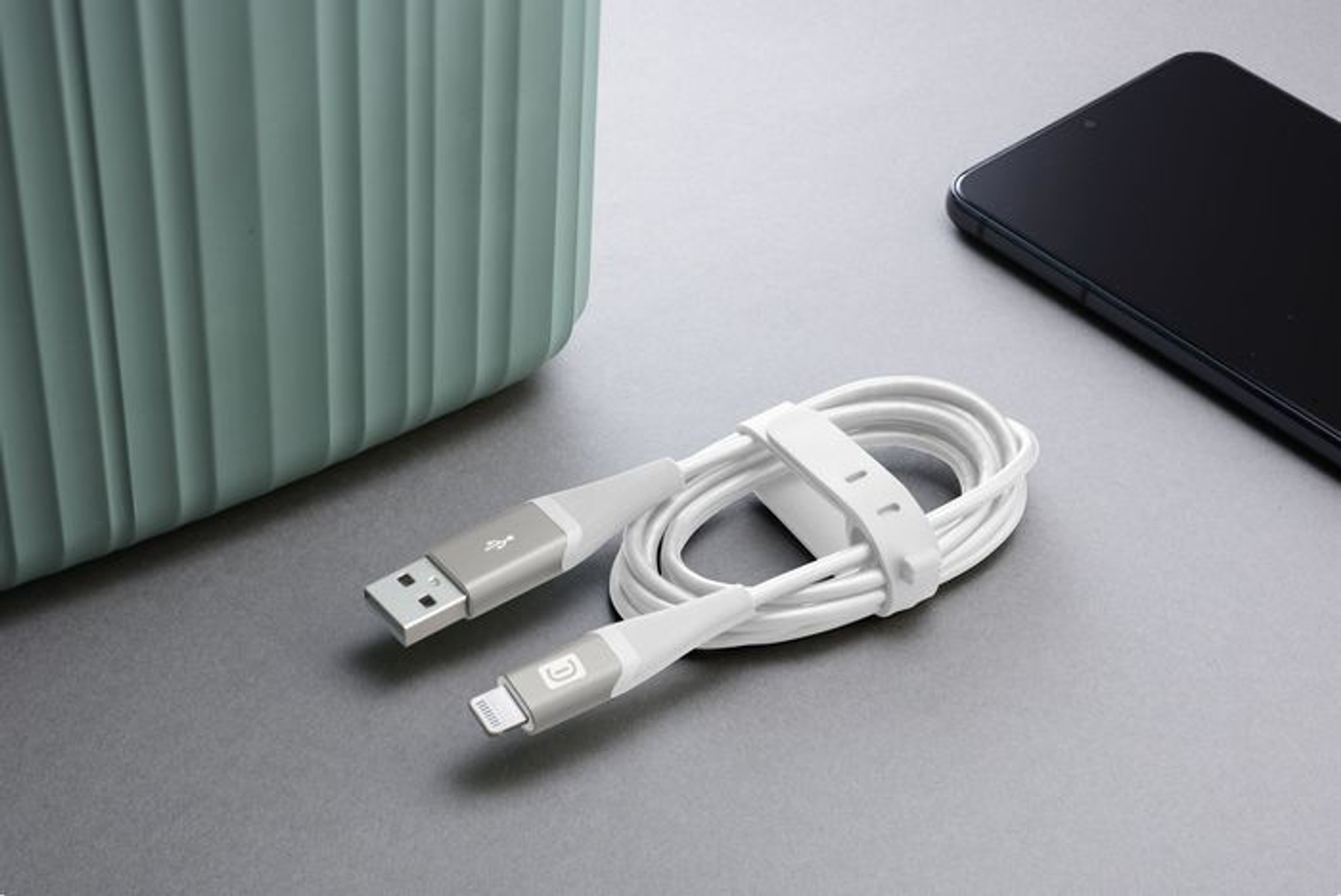 Ein weißes USB-Kabel mit weißem Band neben einem grünen Behälter und einem schwarzen Telefon.