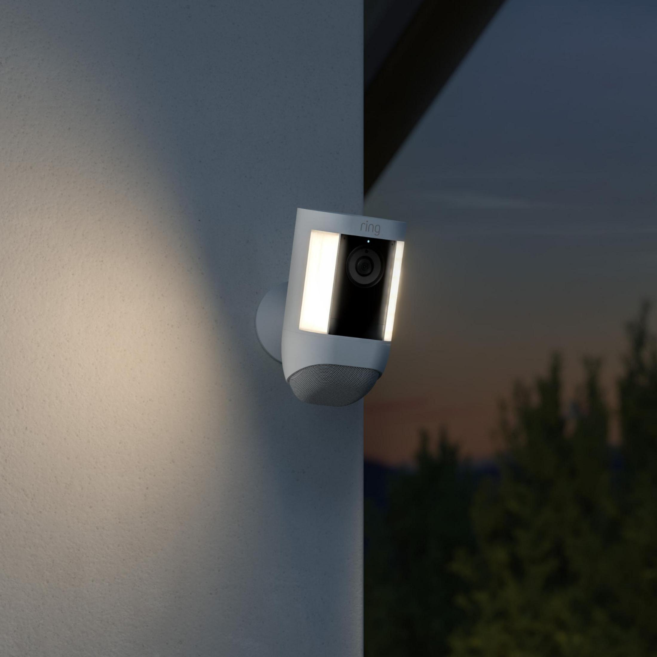 Kamera bezpieczeństwa Ring zamontowana na ścianie, podświetlana nocą. Drzewa w tle.