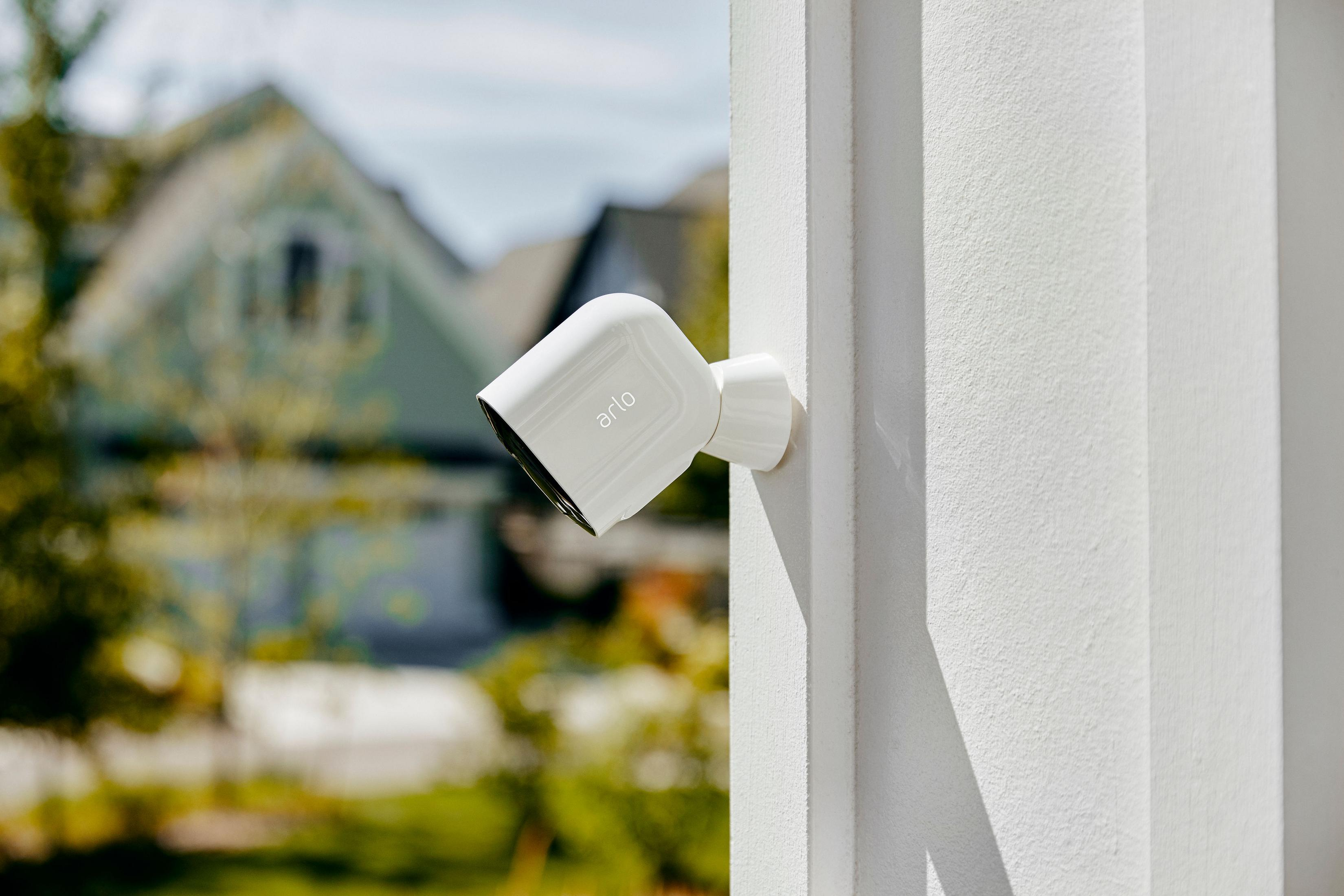 Biała kamera bezpieczeństwa zamontowana na białej ścianie, z rozmytymi domami i drzewami w tle.
