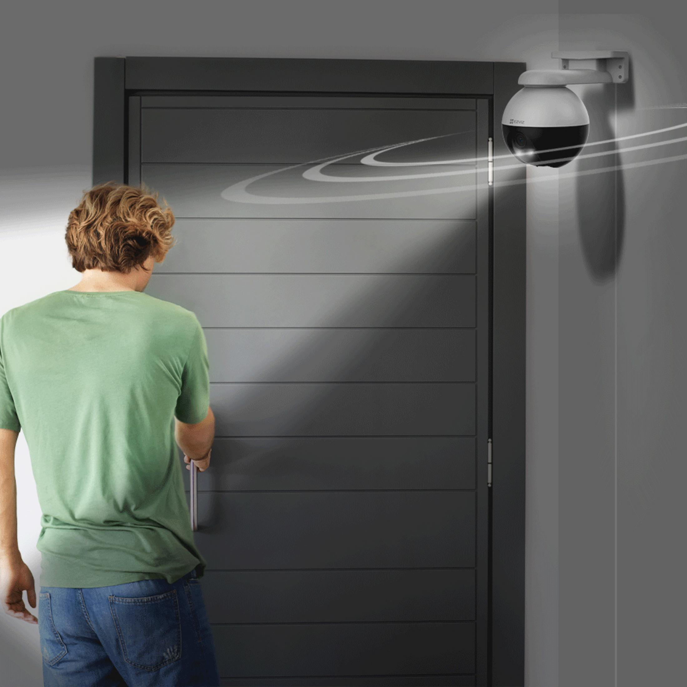 Mężczyzna w zielonej koszuli odblokowuje szare drzwi z kamerą bezpieczeństwa zamontowaną nad nimi. Kamera ma czarny obiektyw.