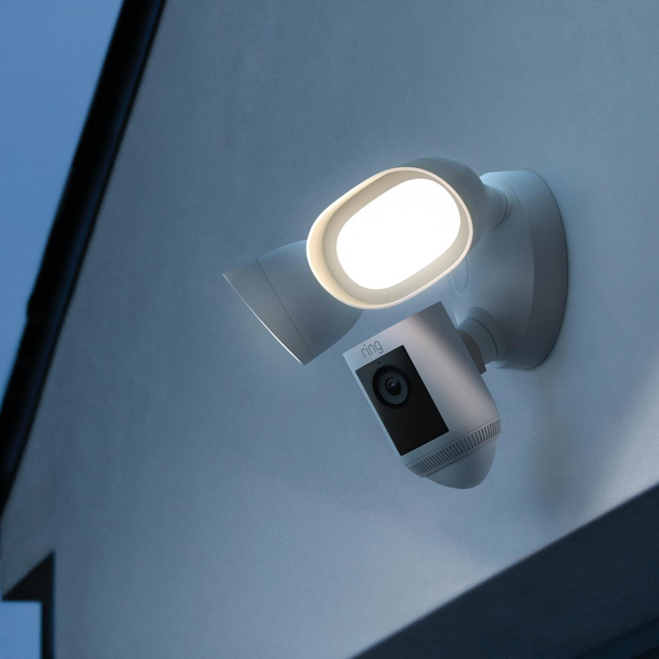 Ein weißes Ring-Sicherheitslicht und eine Kamera, die nachts an einer Wand montiert sind, mit heller Beleuchtung.