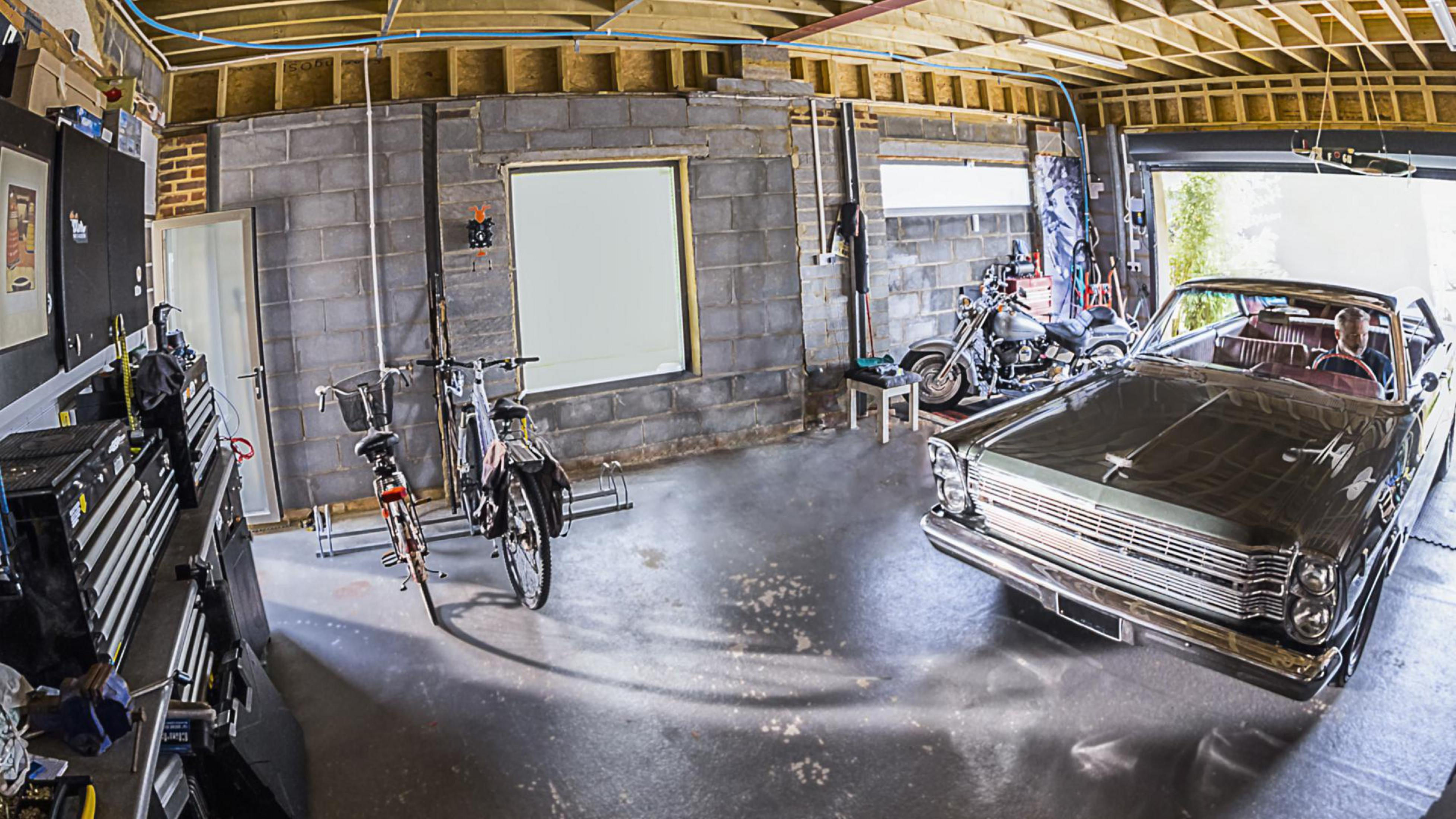 Wnętrze garażu. Widoczne samochód, rowery, narzędzia i skrzynki. Betonowa podłoga, ceglane ściany.
