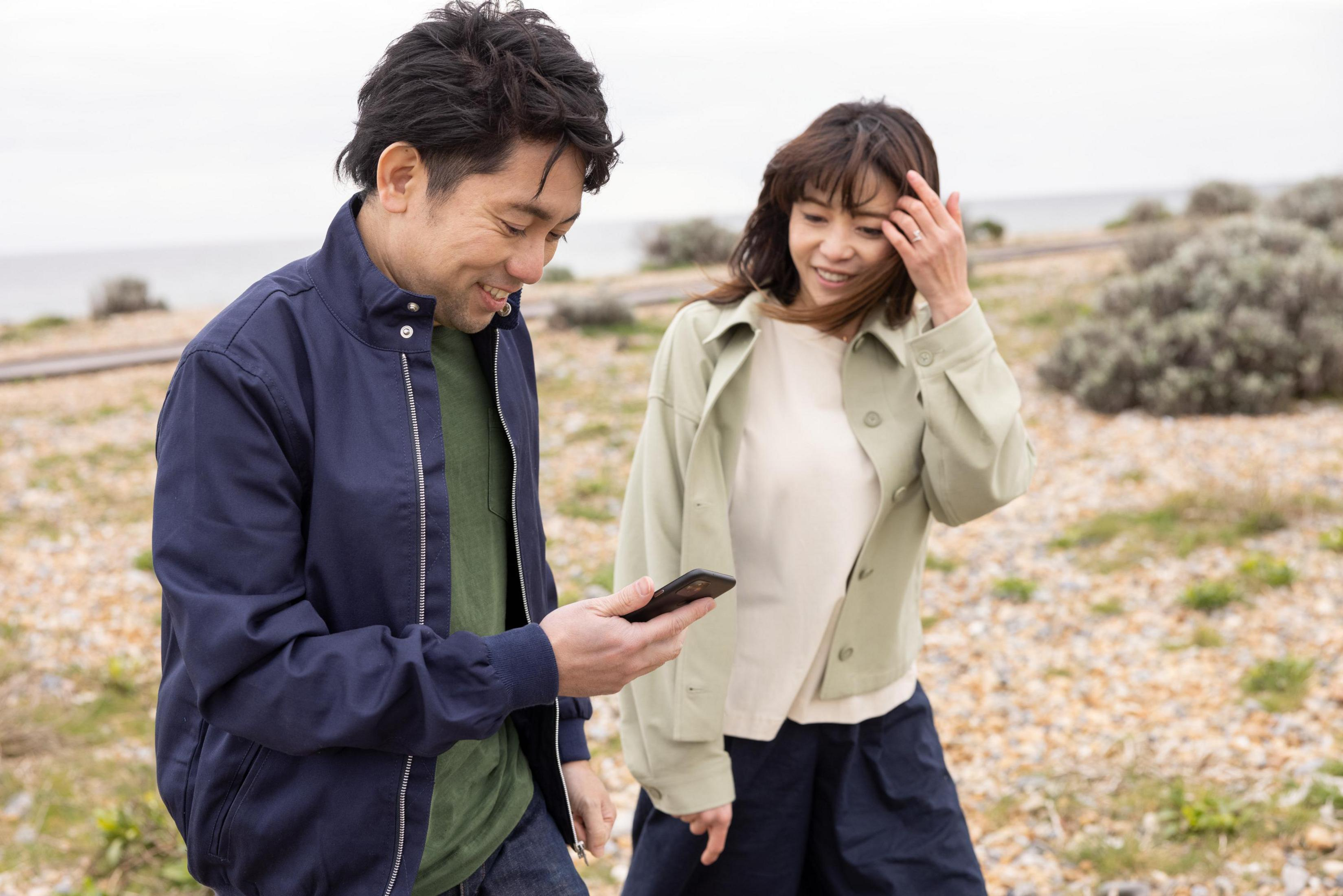 Dwie osoby oglądają telefon na zewnątrz. Mężczyzna w granatowej kurtce, kobieta w jasnozielonej kurtce.