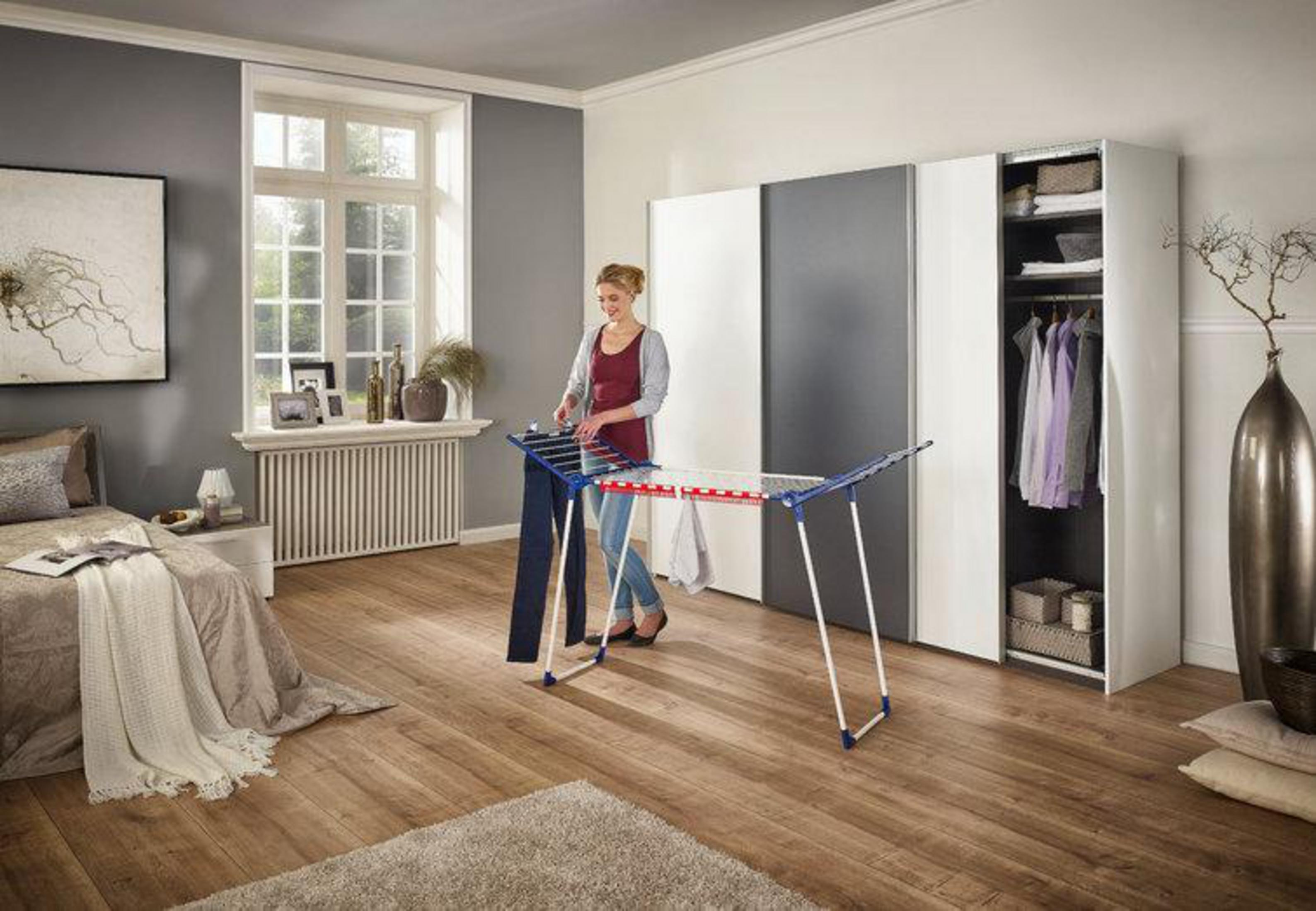 Frau faltet Kleidung auf einem Wäscheständer in einem Schlafzimmer. Schrank und Fenster im Hintergrund.