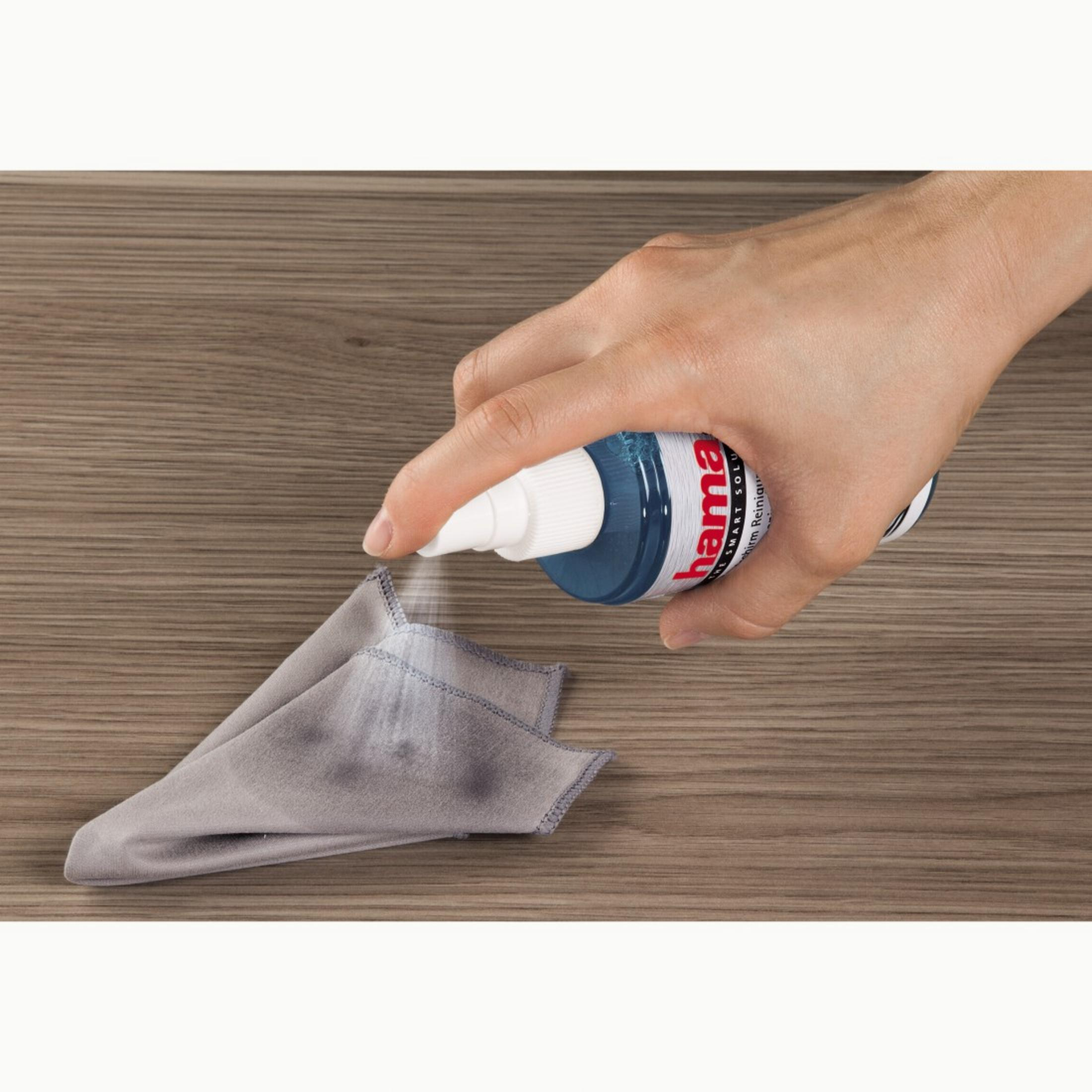 Dłoń spryskująca płyn czyszczący na szarą szmatkę na drewnianym stole. Marka Hama.