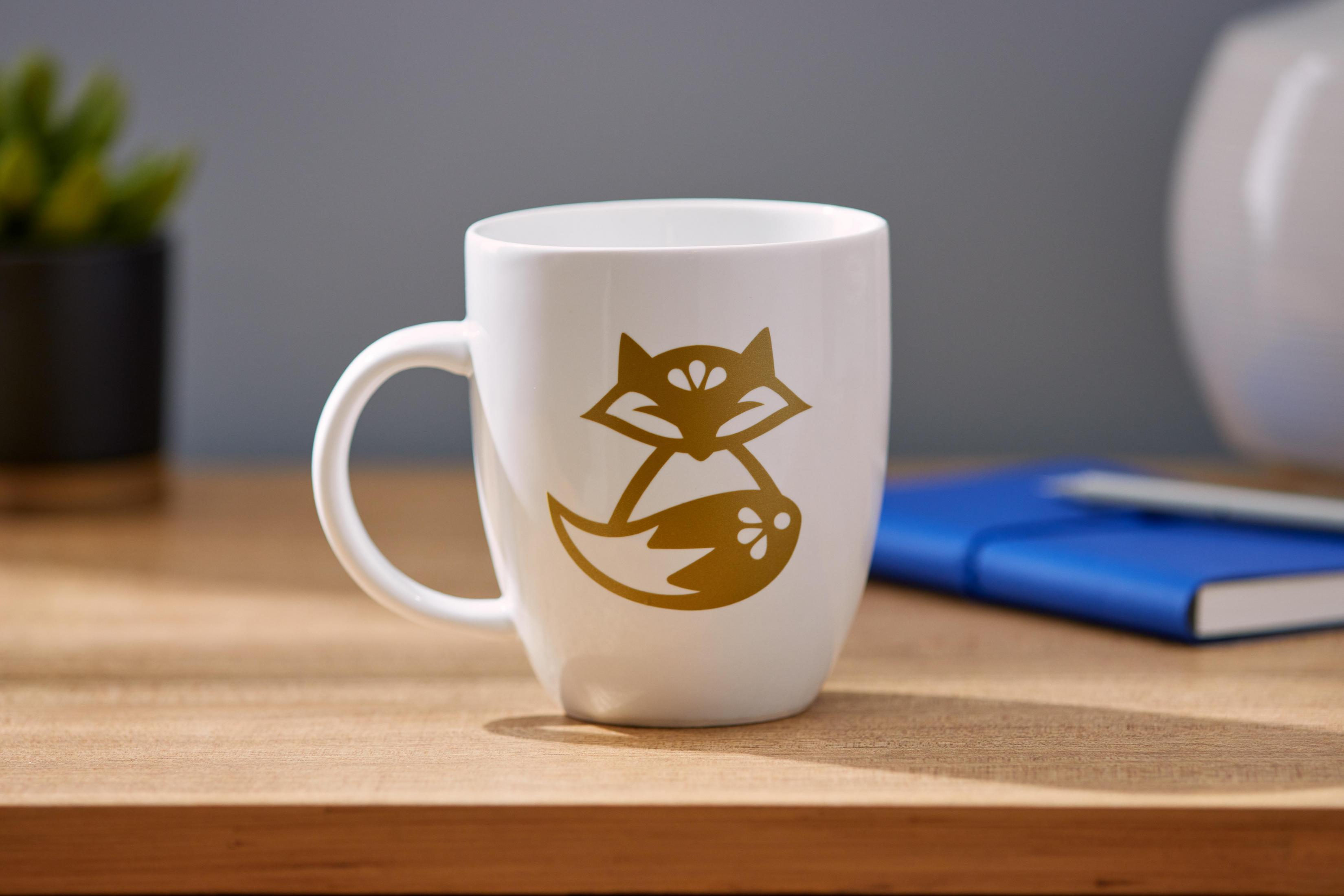 Weißer Becher mit goldenem Fuchsmotiv, auf einem Holztisch.