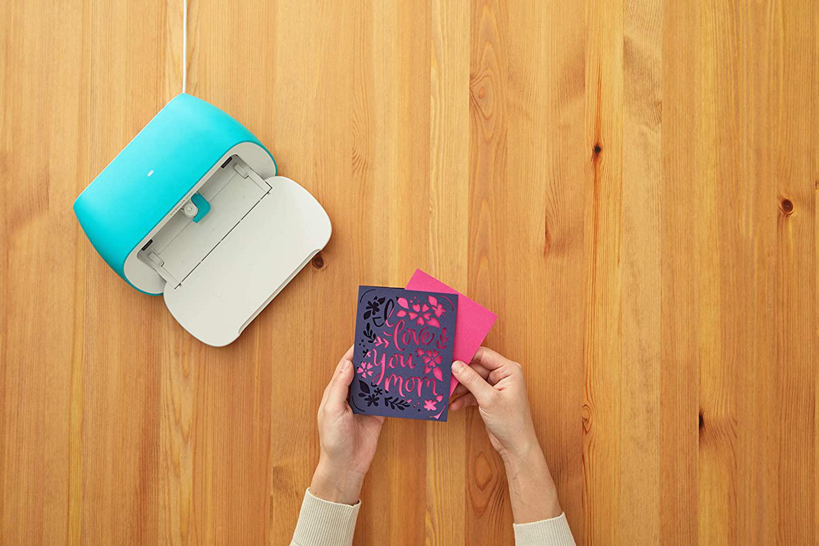 Hände halten Karte mit "Love You Mom", Cricut-Maschine im Hintergrund.