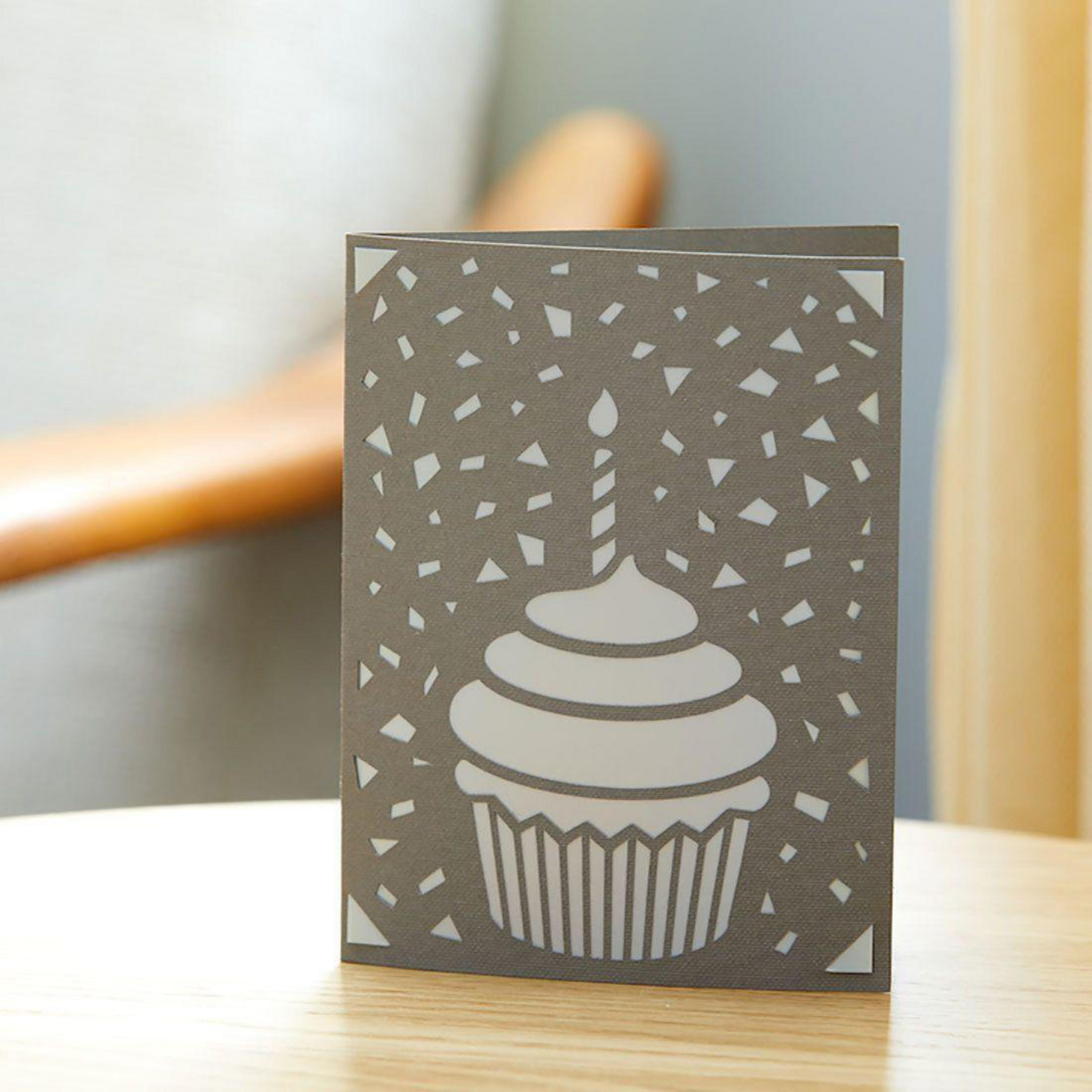 Graue Geburtstagskarte mit Cupcake-Ausschnitt und einer brennenden Kerze. Weißes Konfetti-Design. Tischplatte.