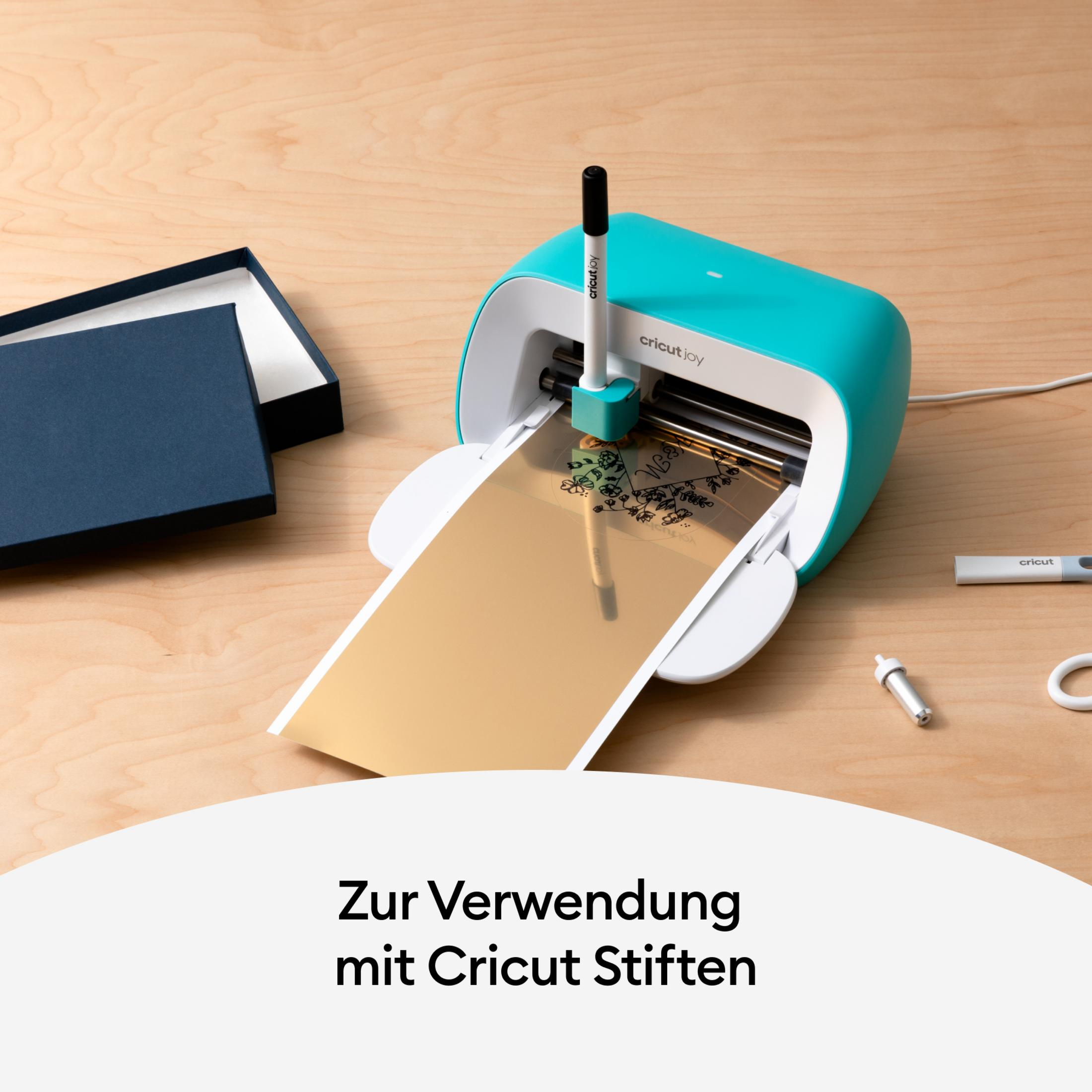 Eine Cricut Joy Maschine, die ein goldenes Material mit einem schwarzen Stift schneidet. Eine blaue Box im Hintergrund.