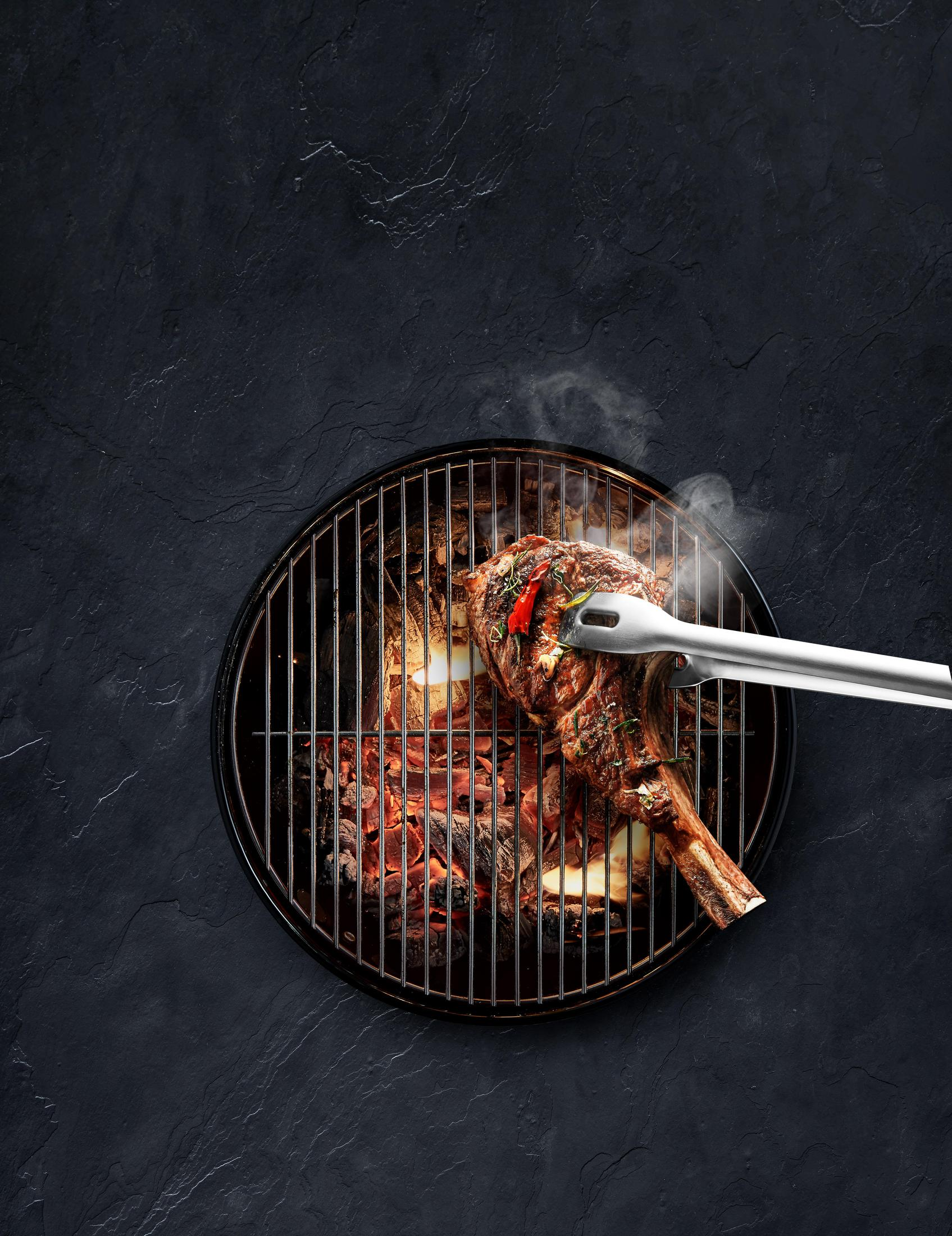 Gegrilltes Steak auf Holzkohlegrill mit Zange, auf dunkler, grauer Steinoberfläche.