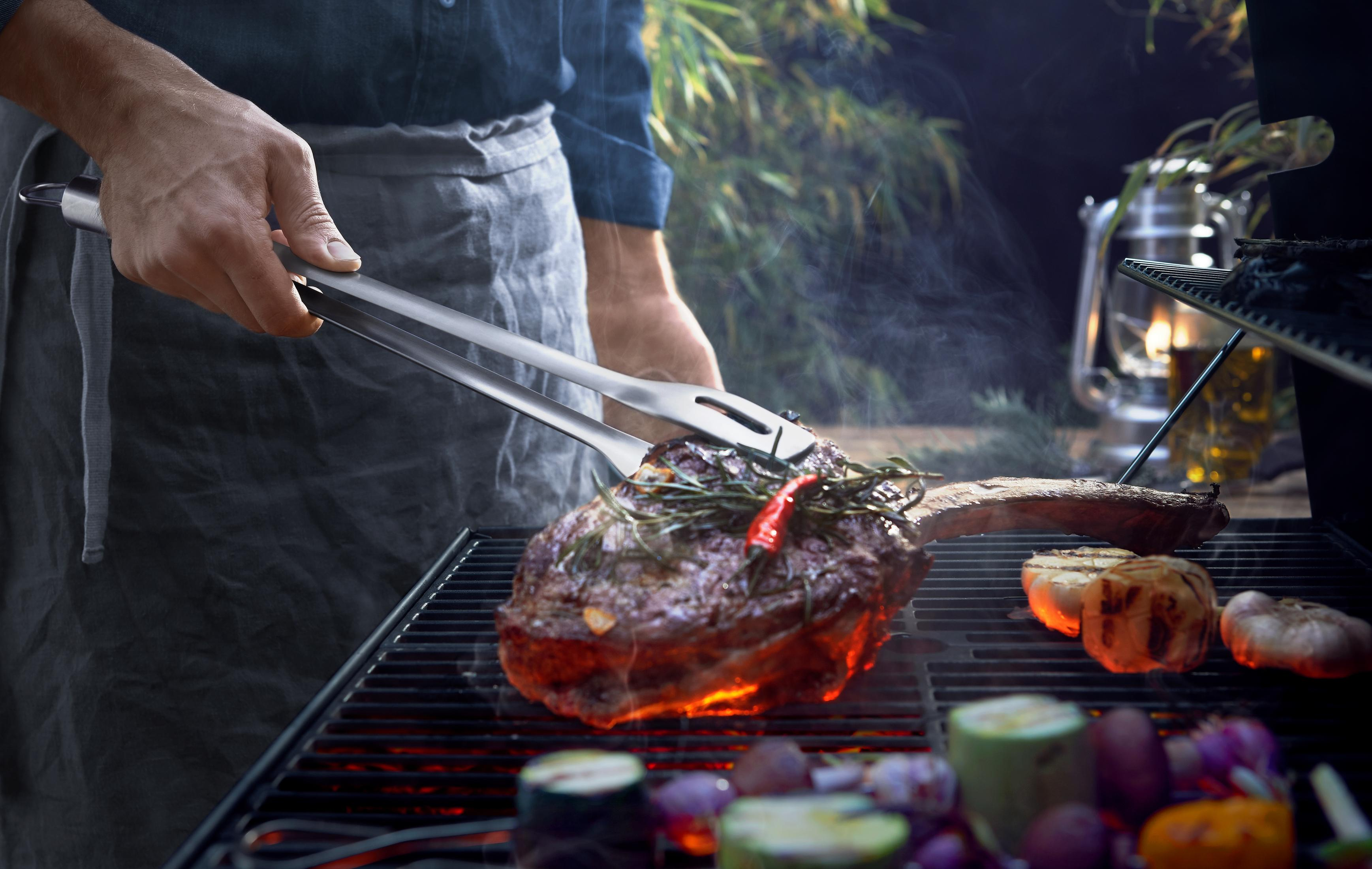 Eine Person grillt ein Steak mit einer Zange auf einem Grill, umgeben von Gemüse und einem Garten im Hintergrund.