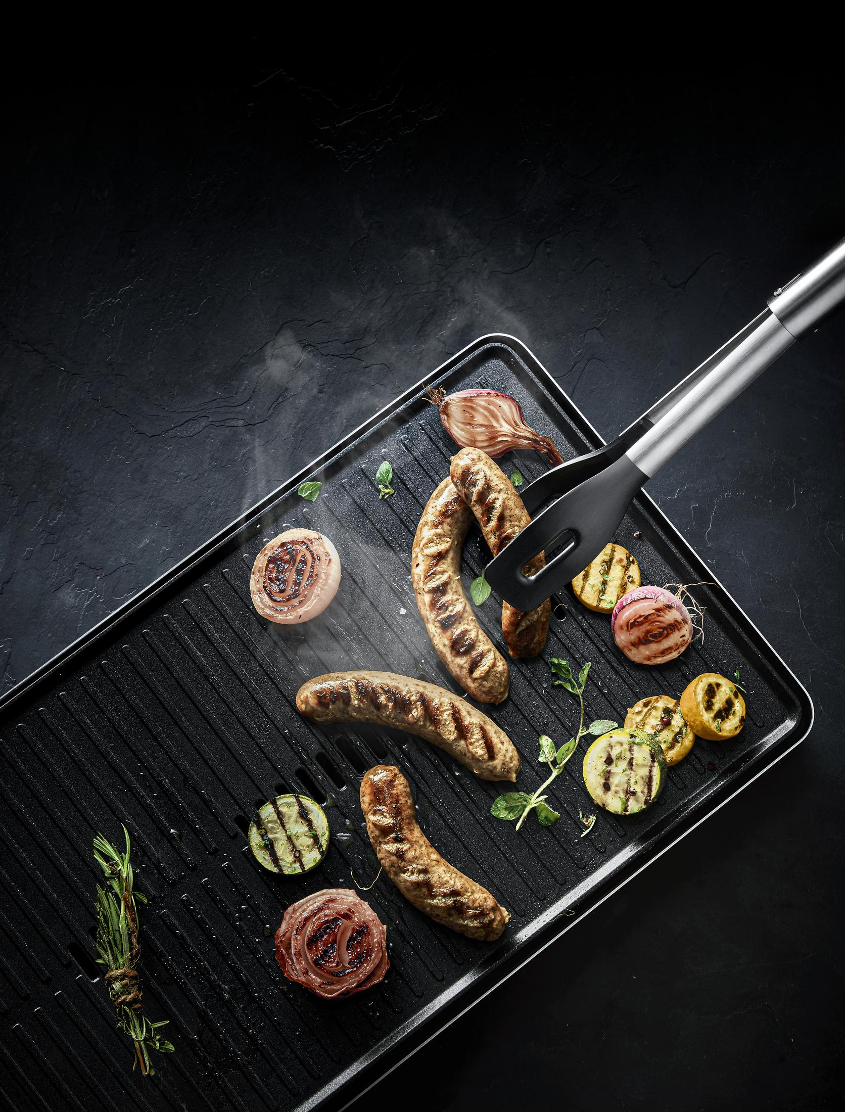 Gegrillte Würstchen und Gemüse auf einem schwarzen Grill. Zangen halten eine Wurst.