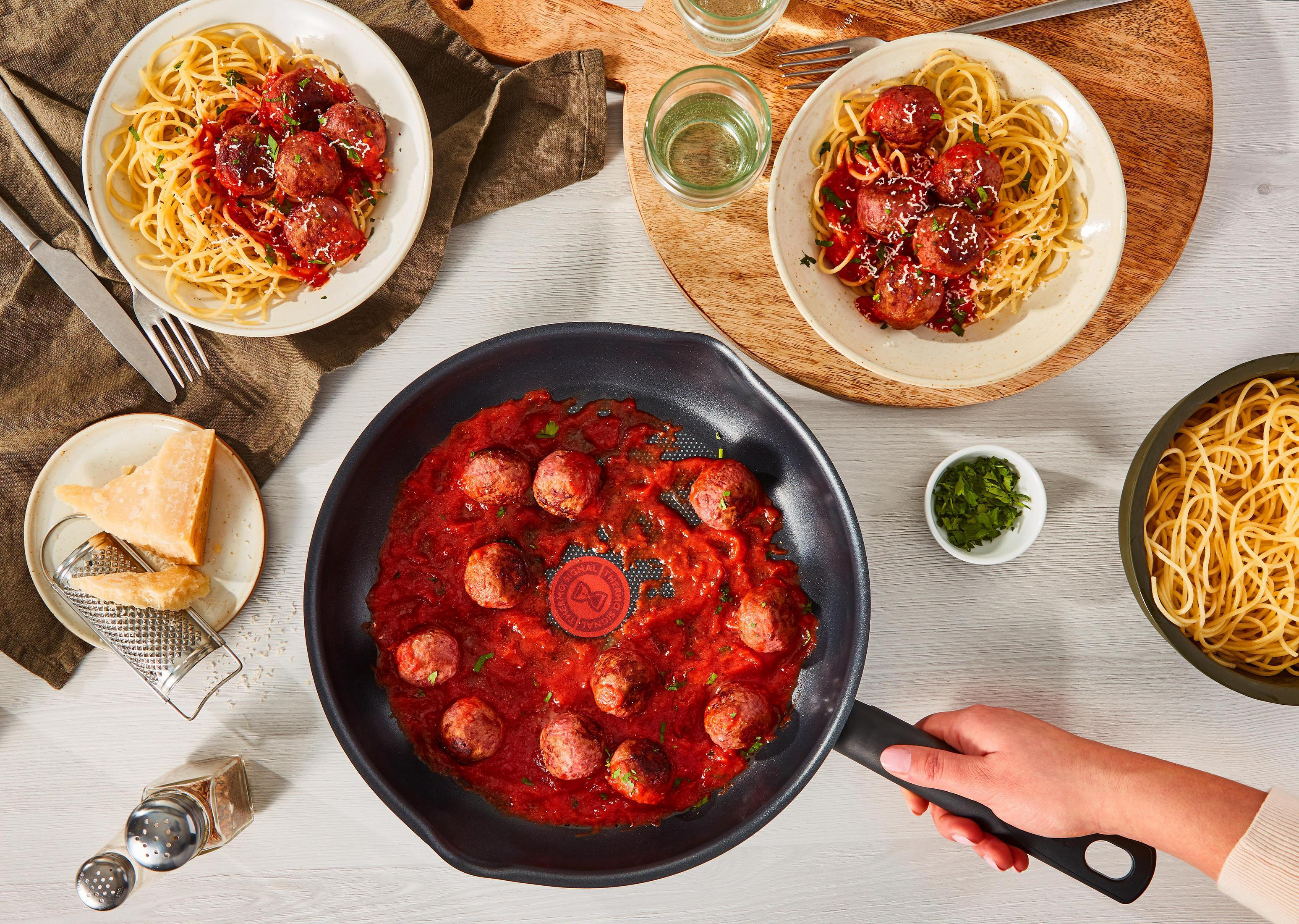 Widok z góry na spaghetti z klopsikami w patelni i na talerzach z parmezanem.