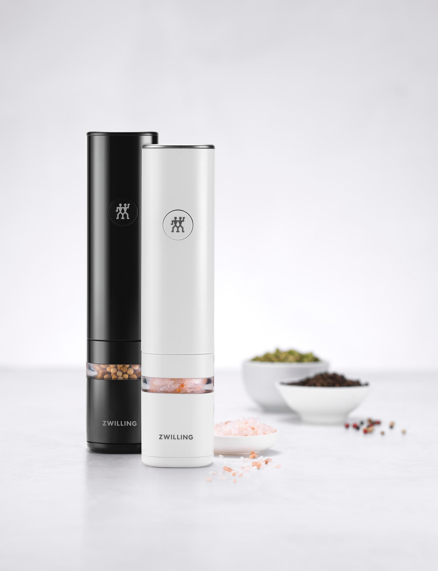 Dwa elektryczne młynki do pieprzu, czarny i biały, z przyprawami w miskach, na białej powierzchni.