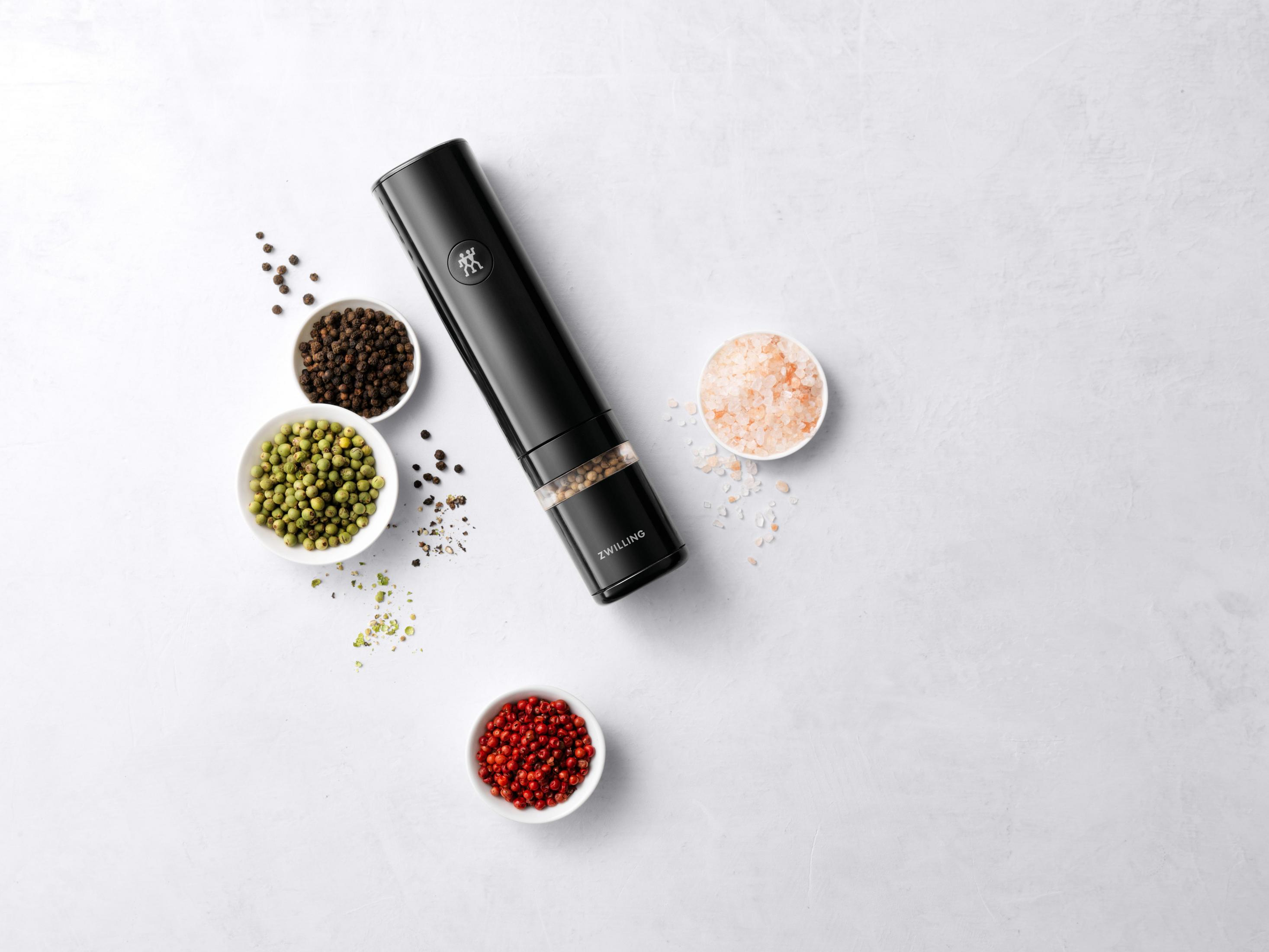 Czarny elektryczny młynek do pieprzu z pieprzem i solą w małych miseczkach na białej powierzchni.