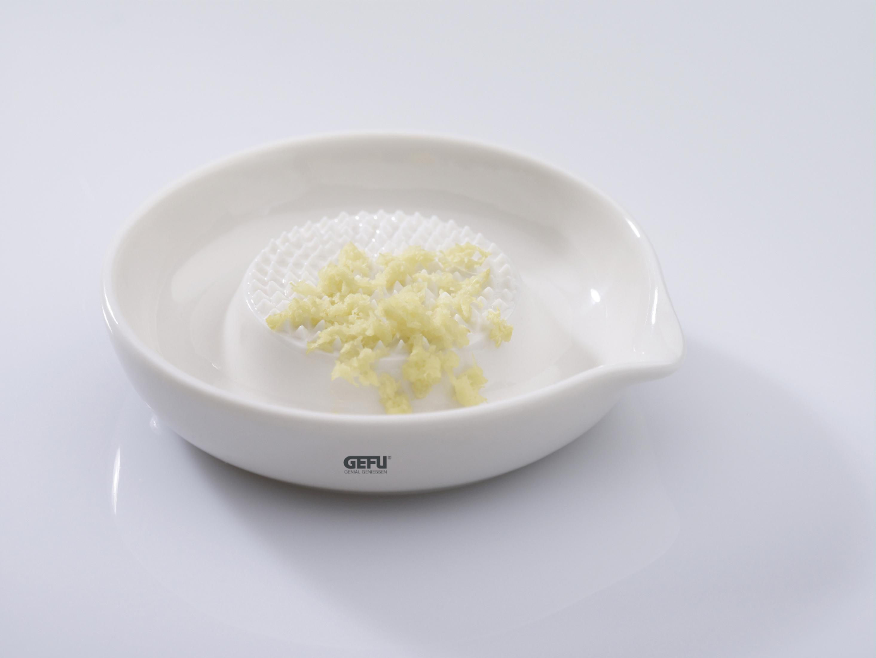 Ein weißer Knoblauchreibe mit geriebenem Knoblauch darauf. Es hat einen Ausguss.