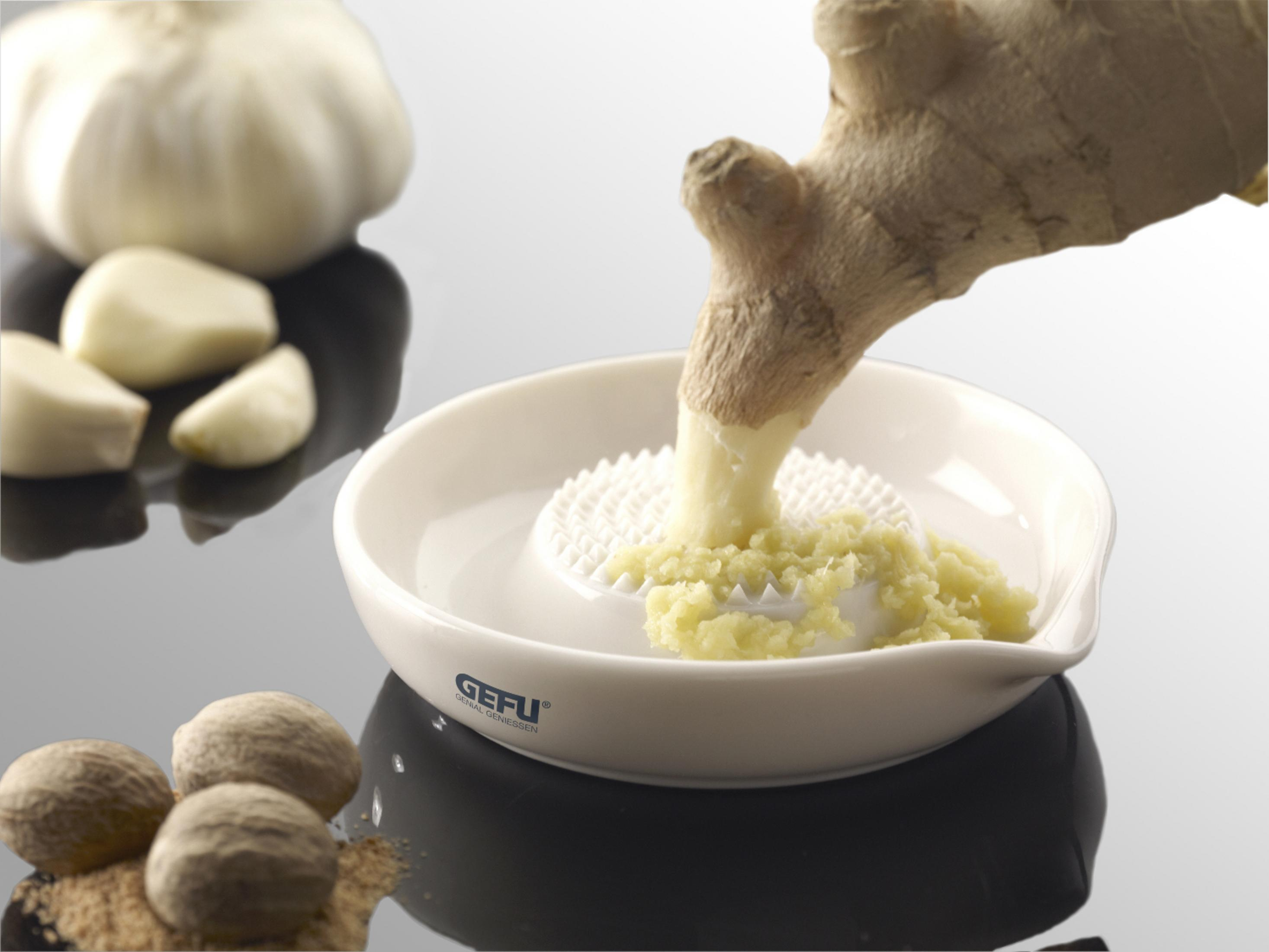 Ingwerwurzel wird in eine weiße Keramikschale gerieben, Knoblauch und Muskatnuss daneben.