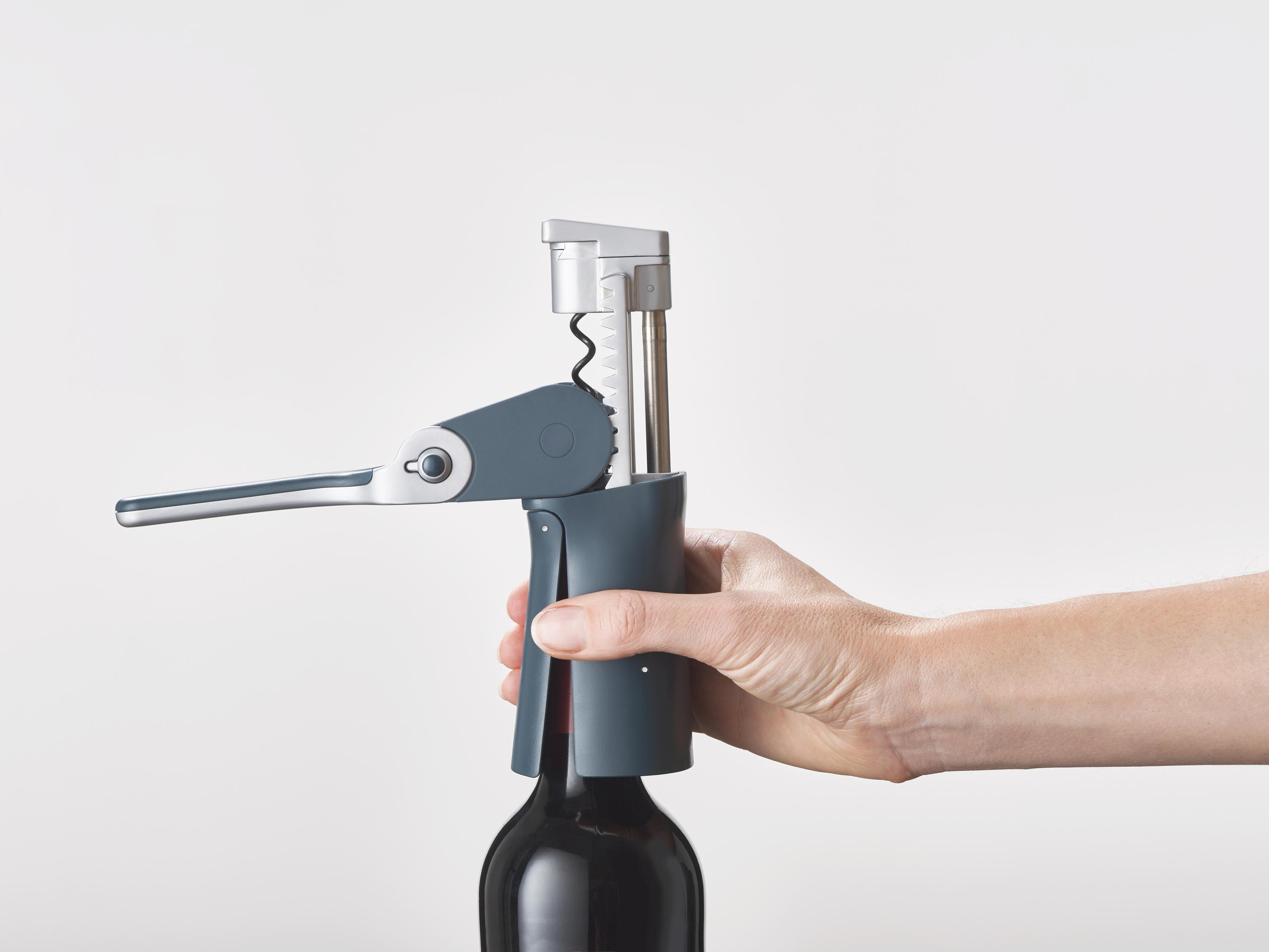 Die Hand einer Person, die einen Weinöffner an einer Weinflasche verwendet. Der Öffner ist grau und silber.