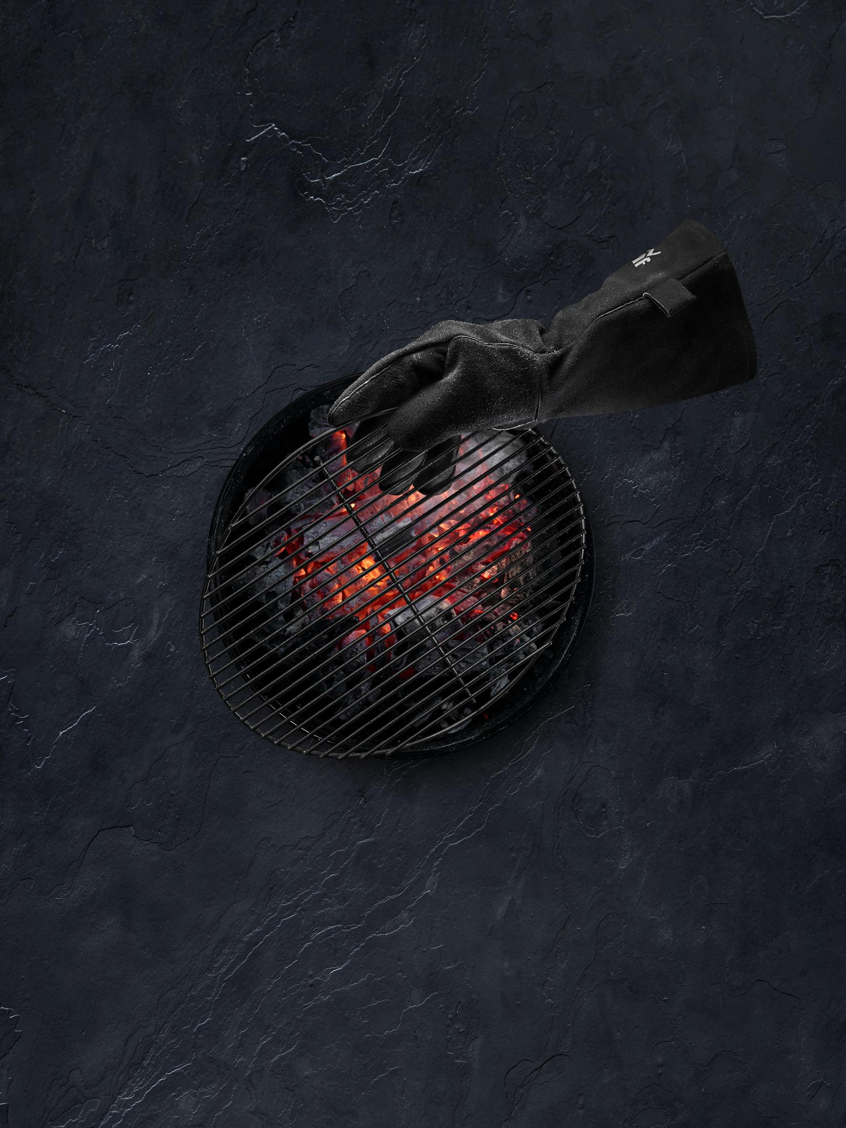 Eine Hand in einem schwarzen Handschuh, die einen Grill mit heißen Kohlen bedient. Dunkler Hintergrund.