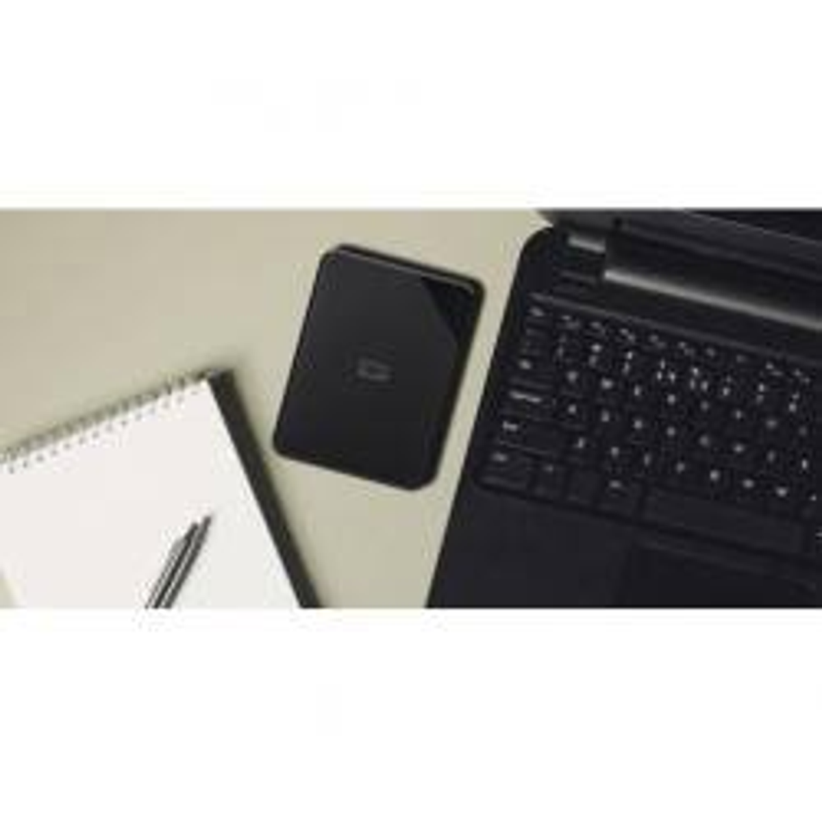 Czarny laptop, czarny zewnętrzny dysk twardy, notatnik i długopis na beżowej powierzchni.
