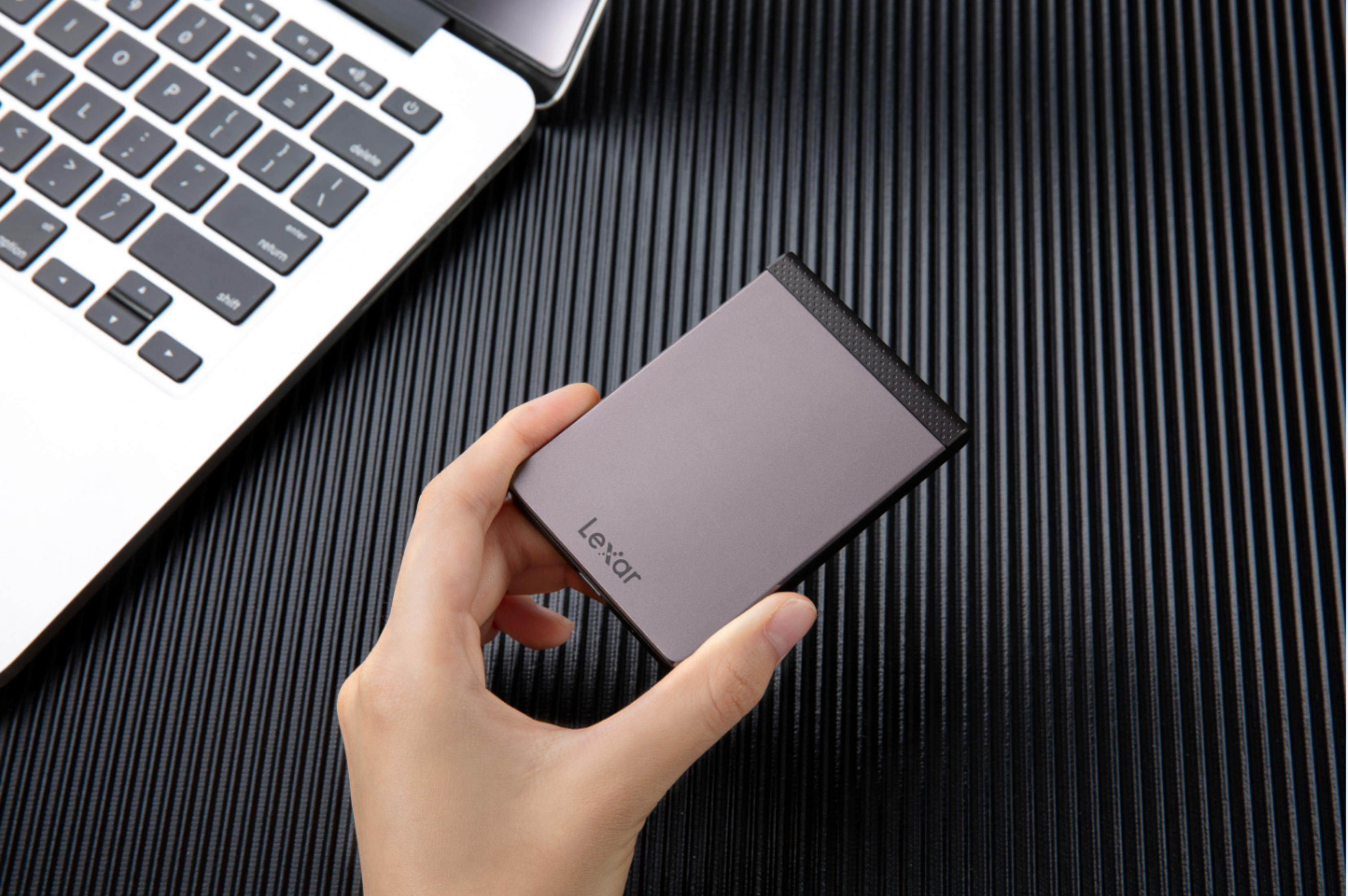 Dłoń osoby trzyma szary zewnętrzny dysk twardy z logo Lexar na czarnej wzorzystej powierzchni.