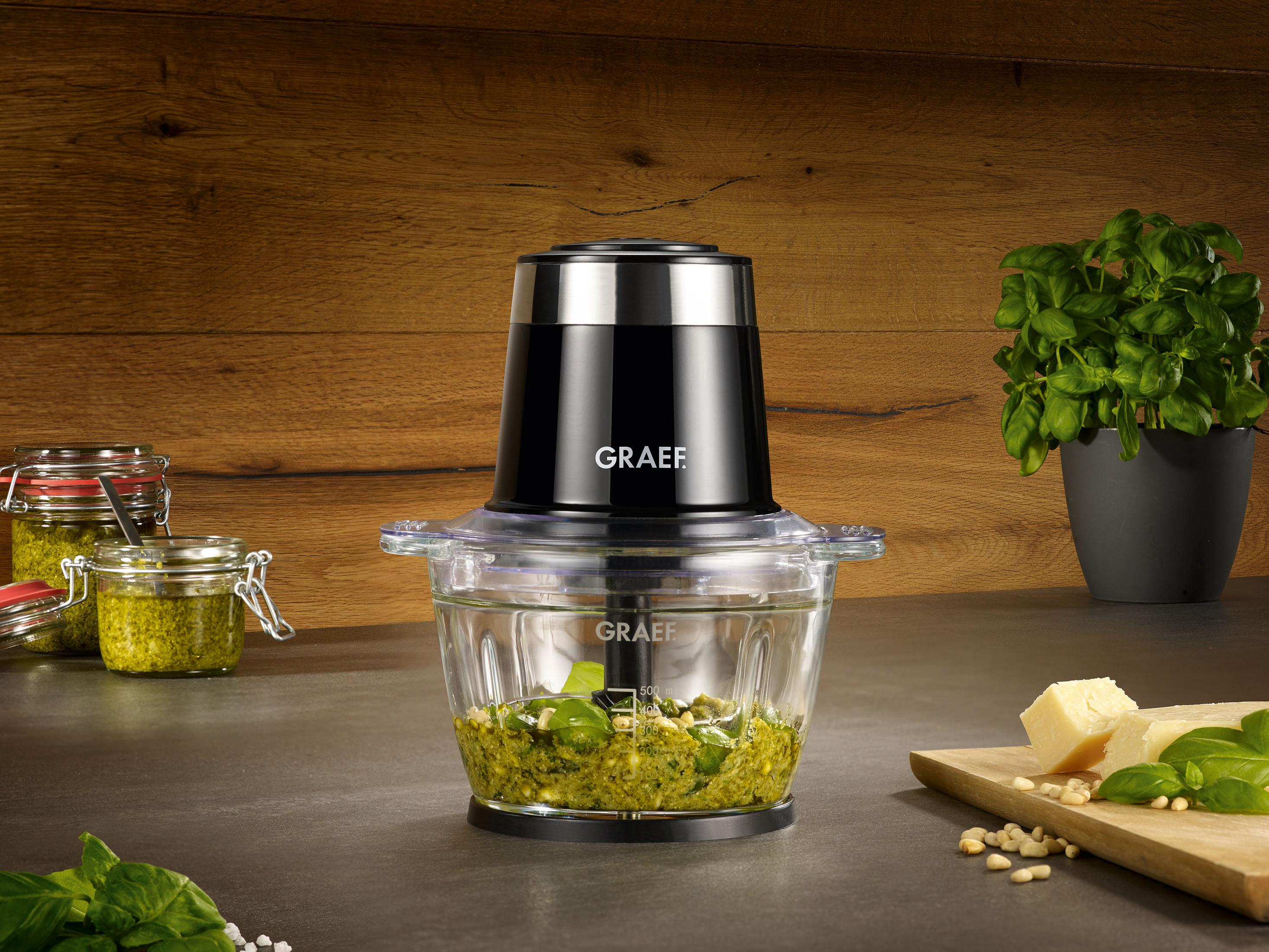 Robot da cucina nero con ciotola in vetro, pesto e ingredienti.