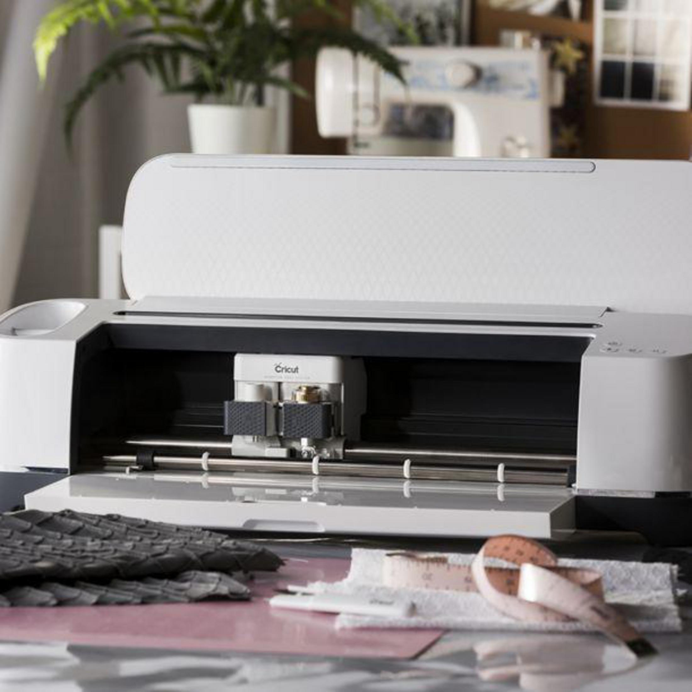 Weiße Cricut-Maschine mit offenem Deckel, umgeben von Bastelmaterialien.