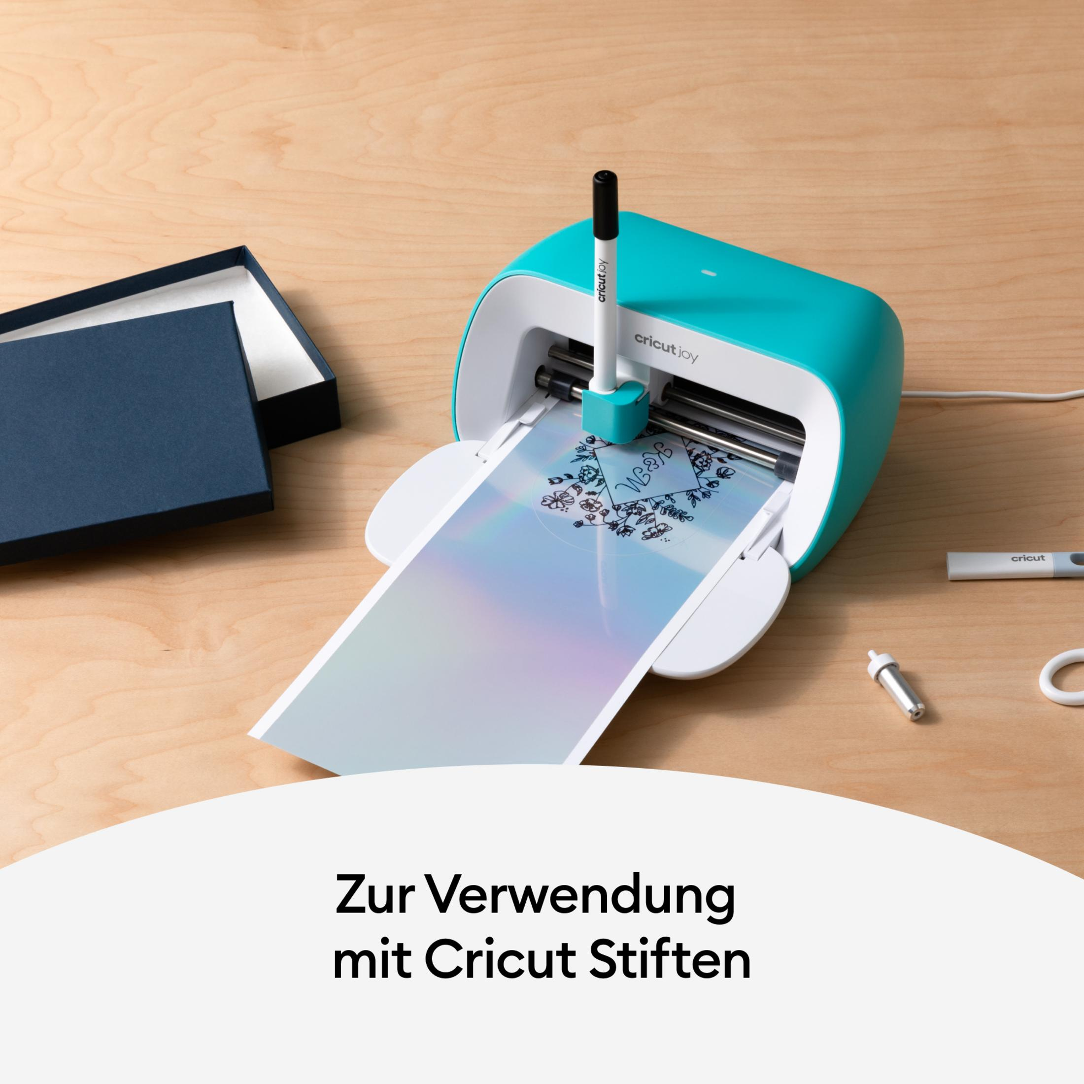 Eine Cricut Joy Maschine mit einem Stift, der auf ein holografisches Blatt druckt. Links ist eine blaue Box.