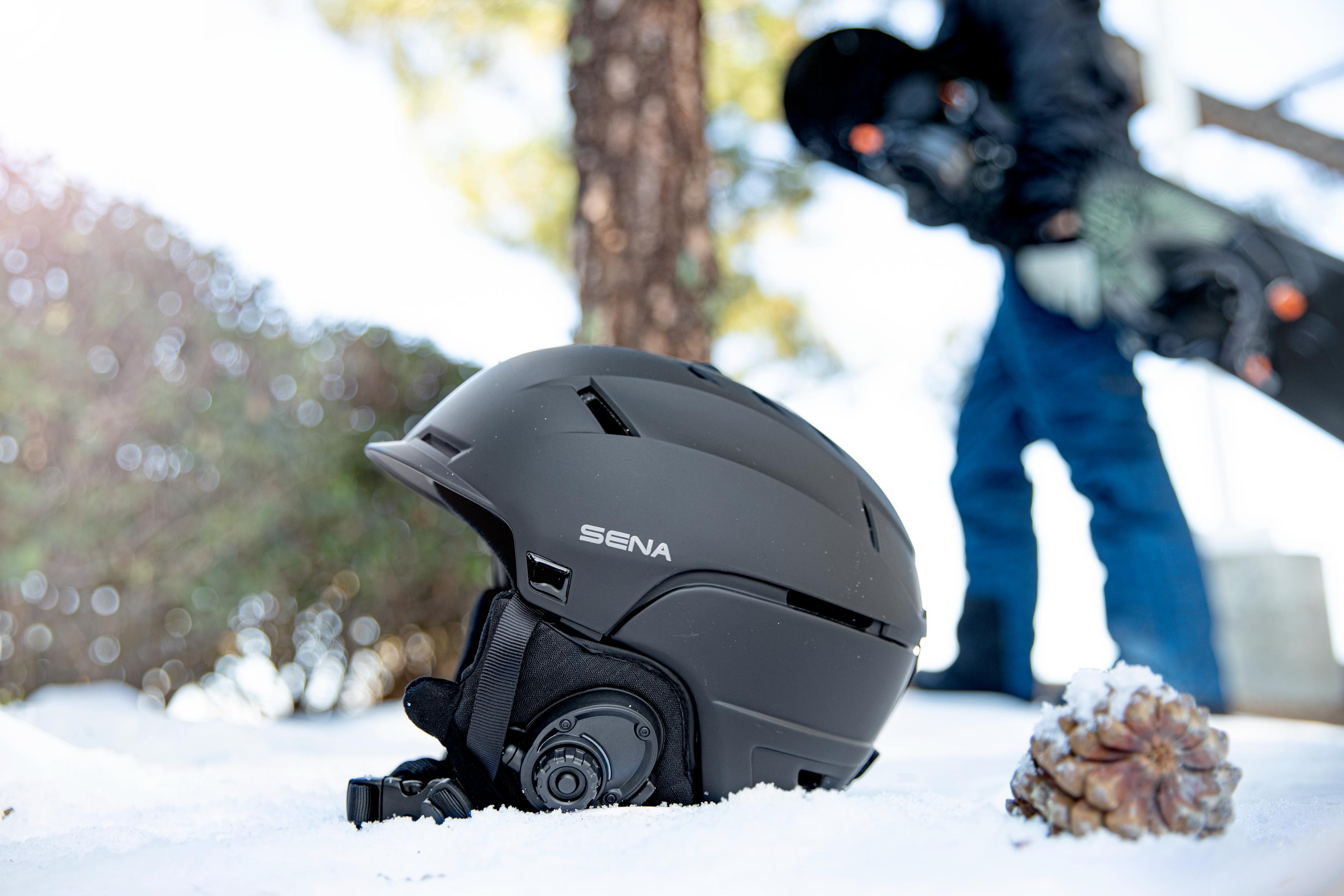 Un casco de esquí negro en la nieve, con una persona y una tabla de snowboard en el fondo.