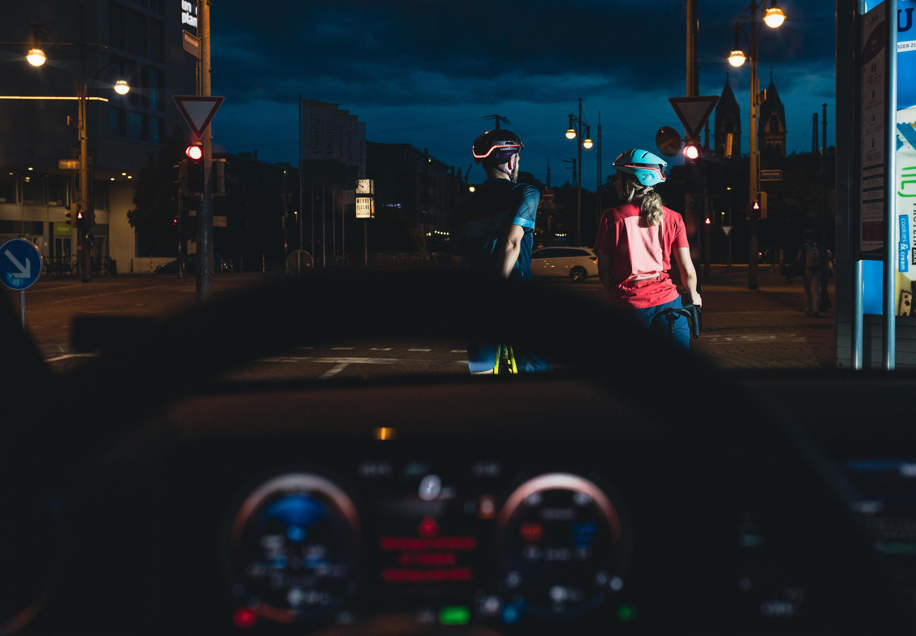 Vista dall'interno di un'auto di due ciclisti su un attraversamento pedonale notturno. Luci della città e cielo scuro.