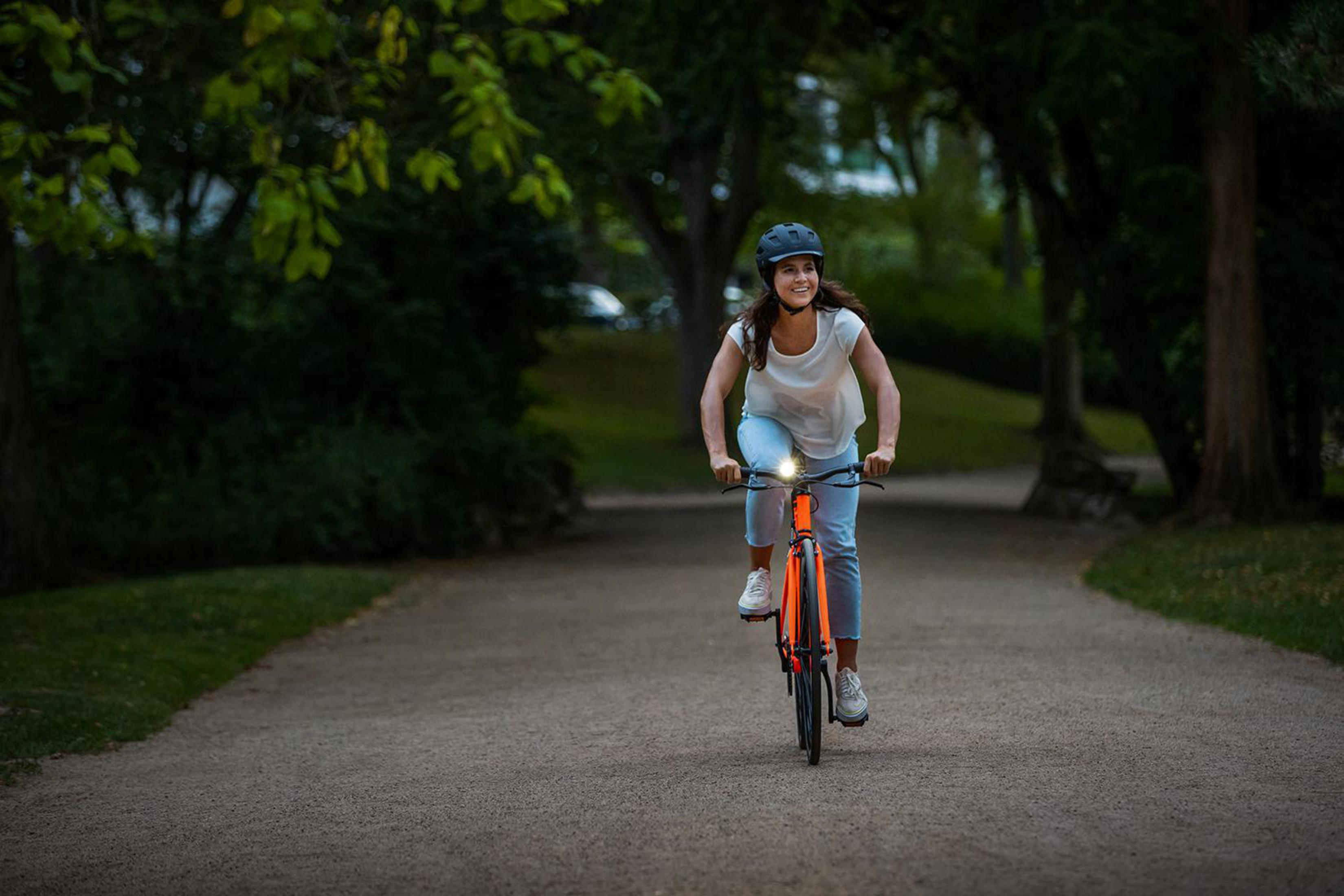 Kobieta jadąca na rowerze ścieżką w parku. Ma na sobie kask i się uśmiecha.