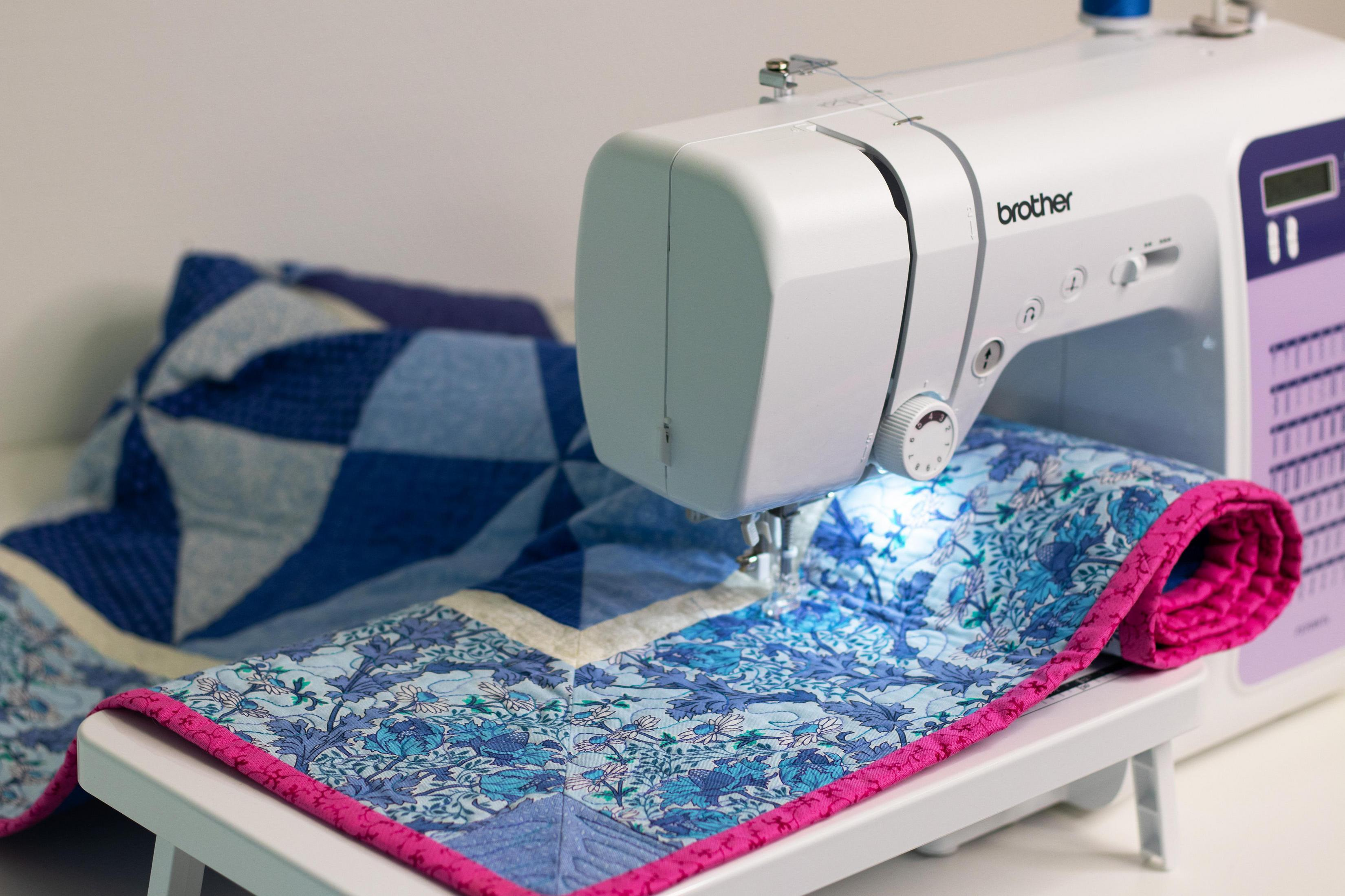 Biała maszyna do szycia szyjąca niebieską patchworkową kołdrę z różowym wykończeniem.