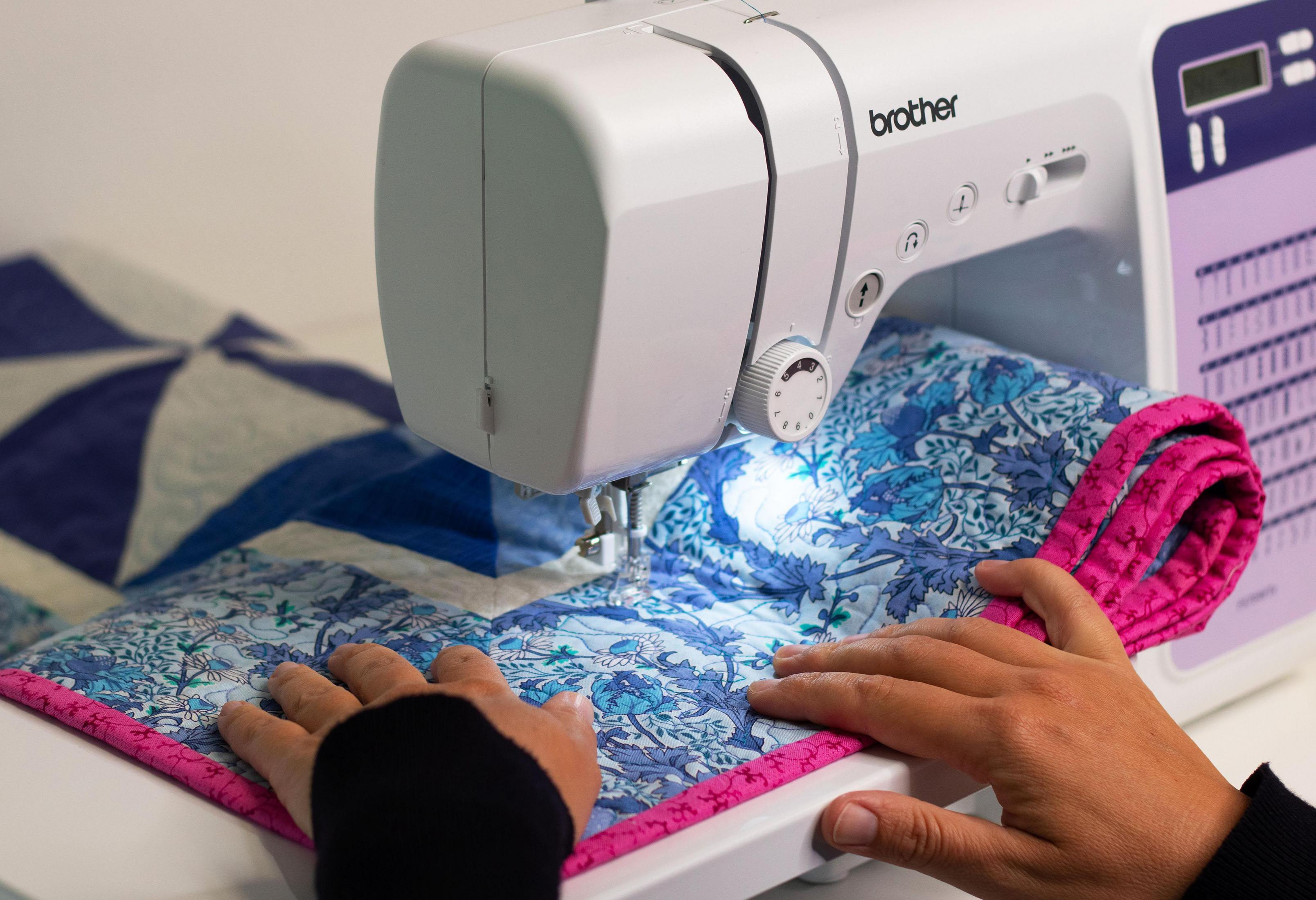 Ręce szyjące niebiesko-białą kwiecistą kołdrę z różową obwódką za pomocą maszyny do szycia Brother.