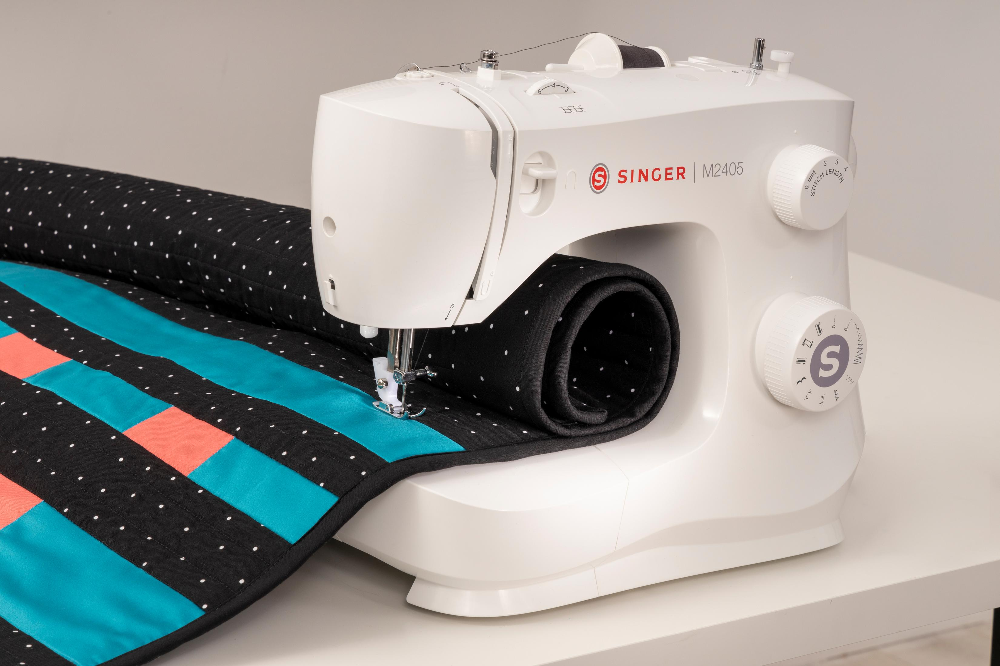 Biała maszyna do szycia Singer szyjąca kołdrę z czarnej tkaniny i kolorowych kwadratów.