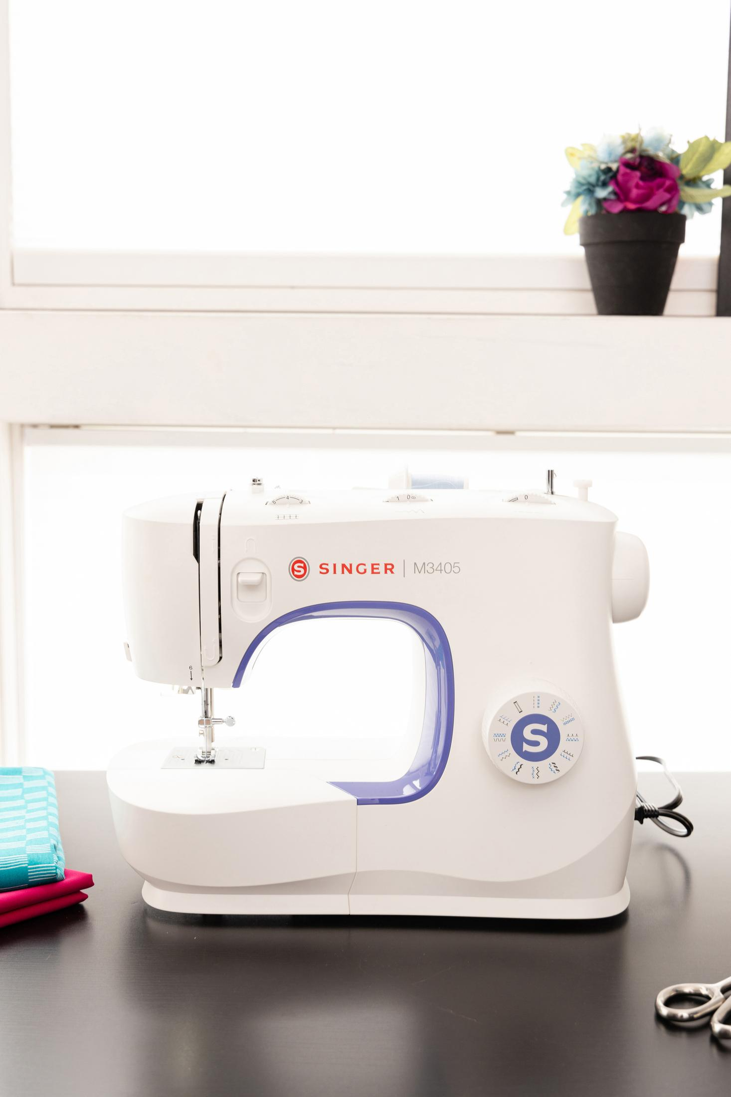 Biała maszyna do szycia na czarnym stole, w tle okno i kwiaty.