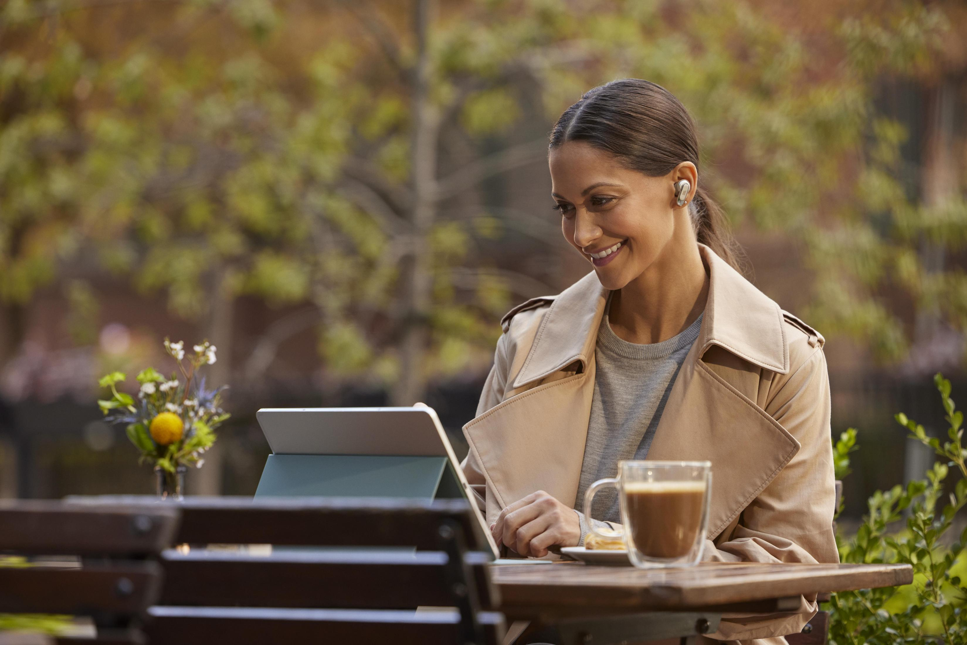 Donna che usa un tablet al tavolo di un bar, con caffè e piatto, alberi sullo sfondo.