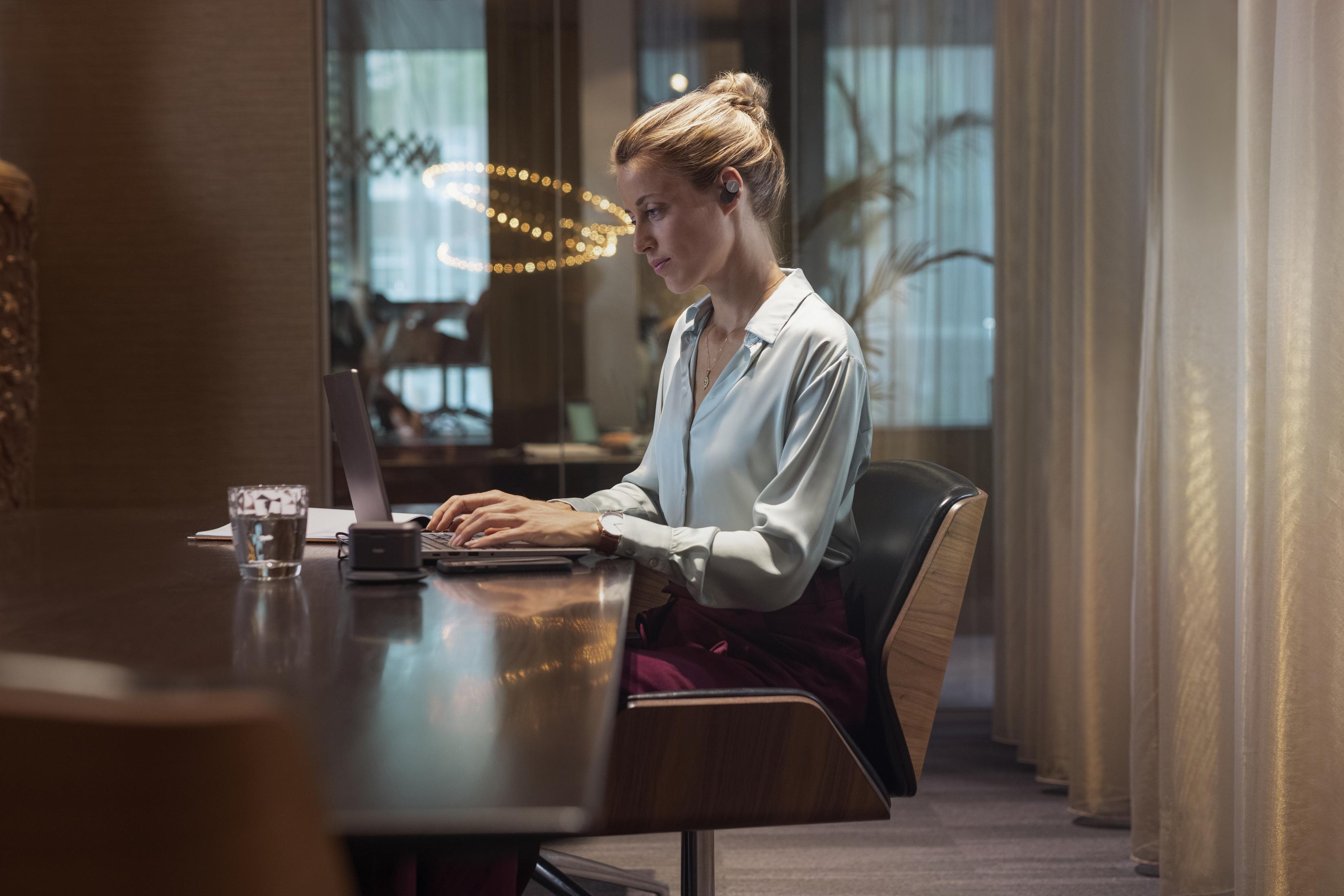 Frau in einem Sitzungssaal mit Laptop. Sie hat Ohrhörer drin. Ein Glas Wasser ist sichtbar.