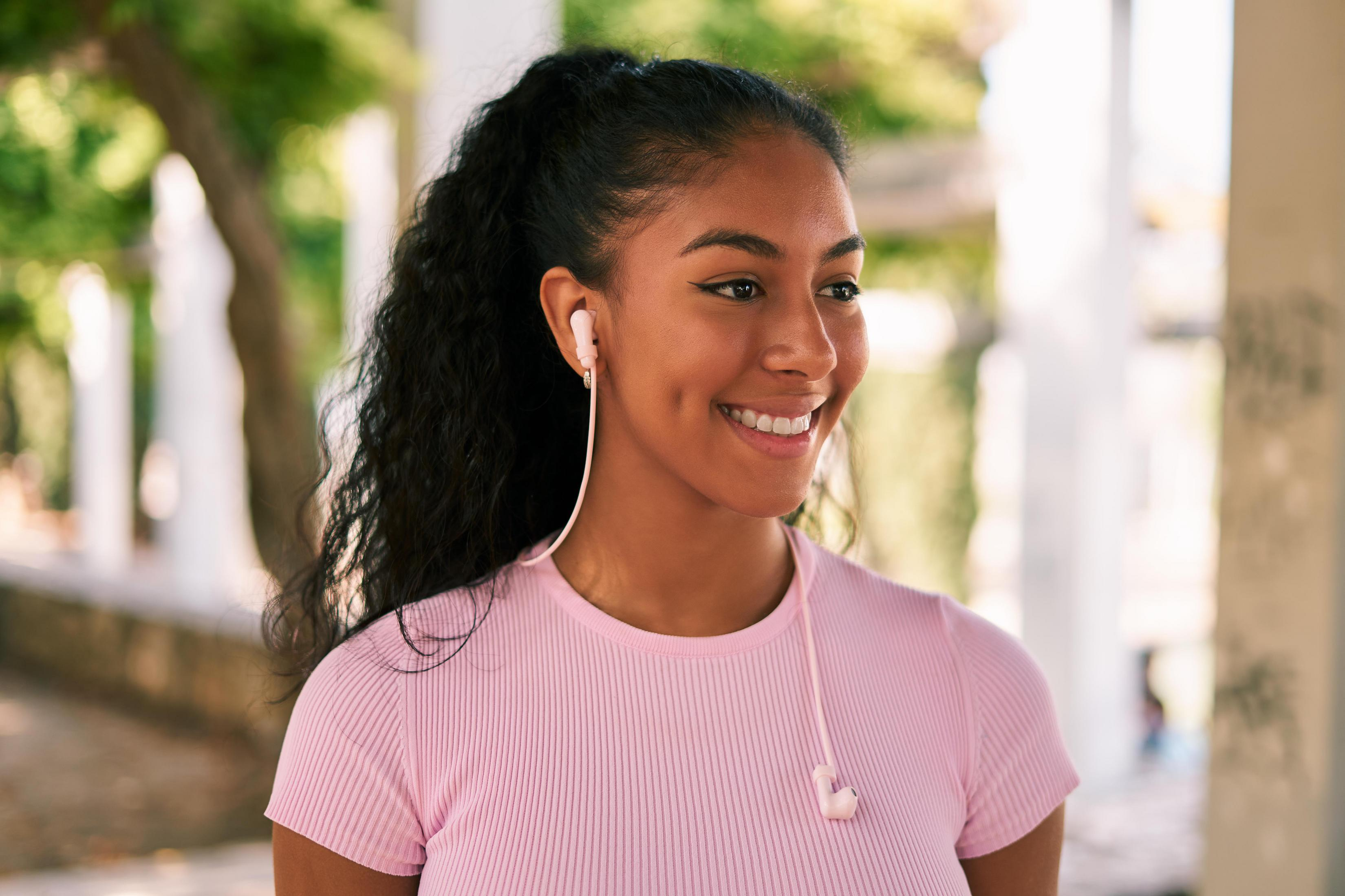 Woman with curly hair smiling, wearing pink shirt and earbuds. Trees in the background.